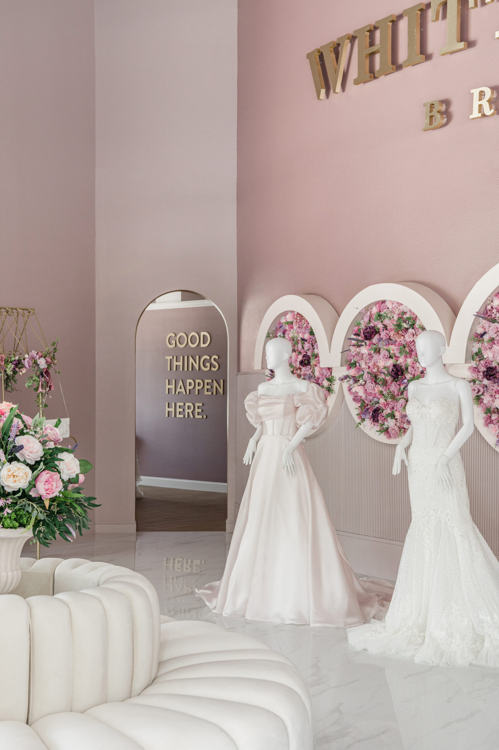 Mannequins dressed in bridal gowns on display in a bridal boutique, with a floral backdrop and an archway with a sign reading "Good Things Happen Here."