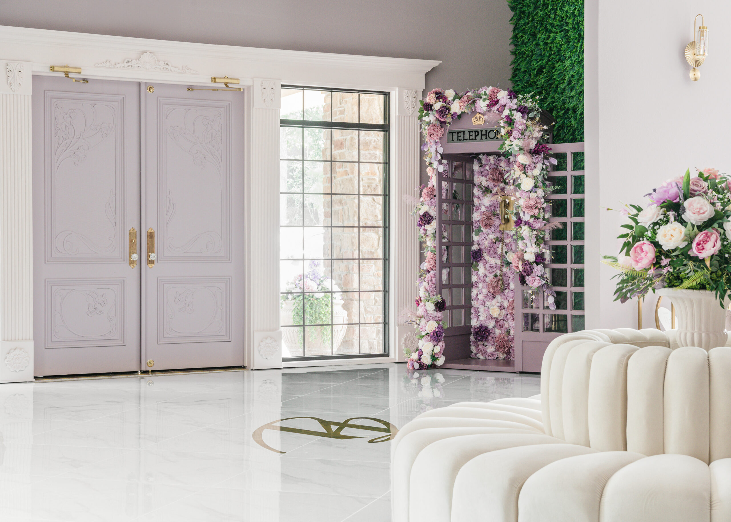 A beautifully decorated entrance area of a bridal boutique, featuring a vintage telephone booth adorned with a floral arrangement. Large glass doors provide a view of the outside greenery.