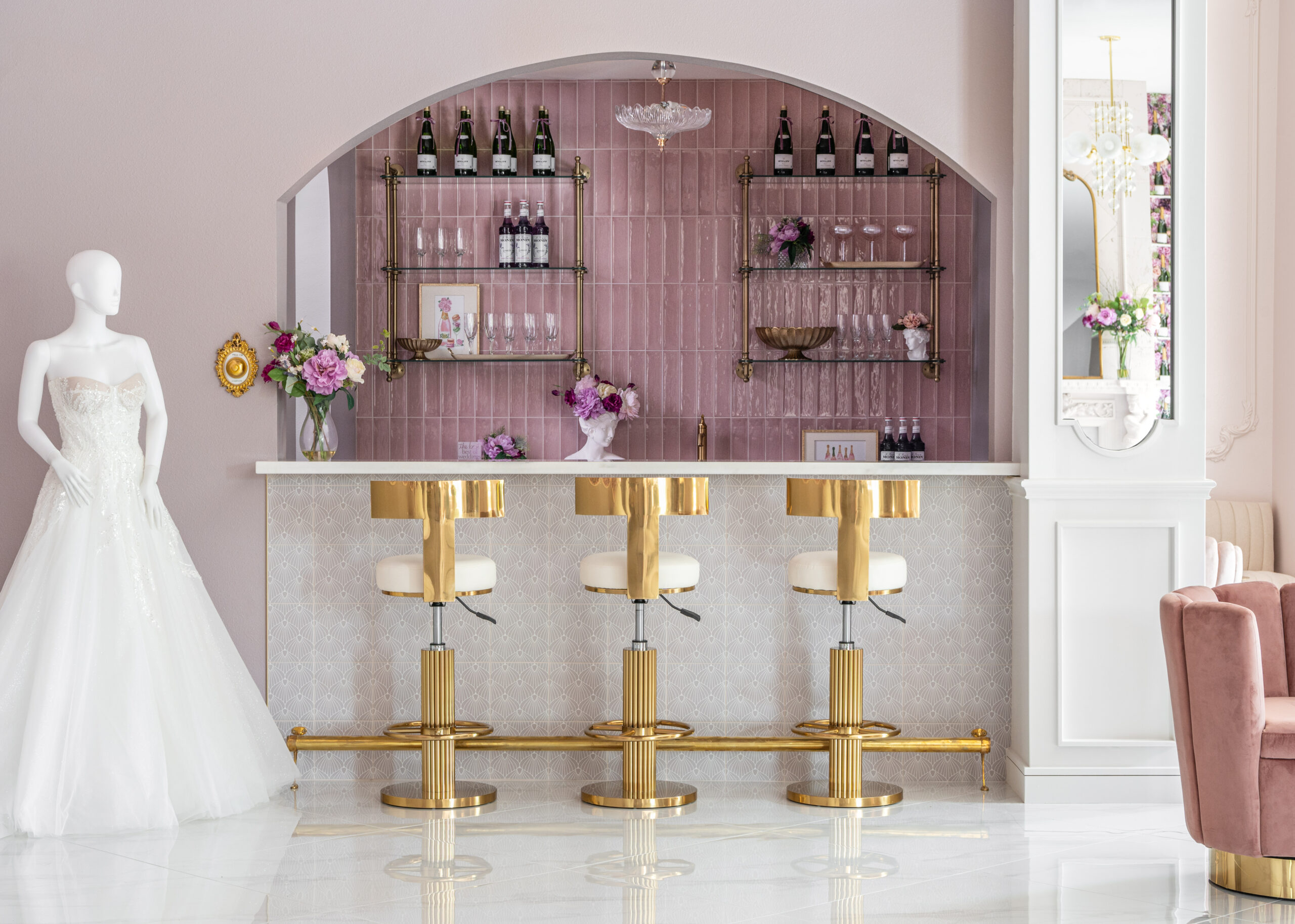 An elegant bar area in a bridal boutique with gold stools, a white countertop, and shelves with glasses, bottles, and floral decor against a pink-tiled wall.