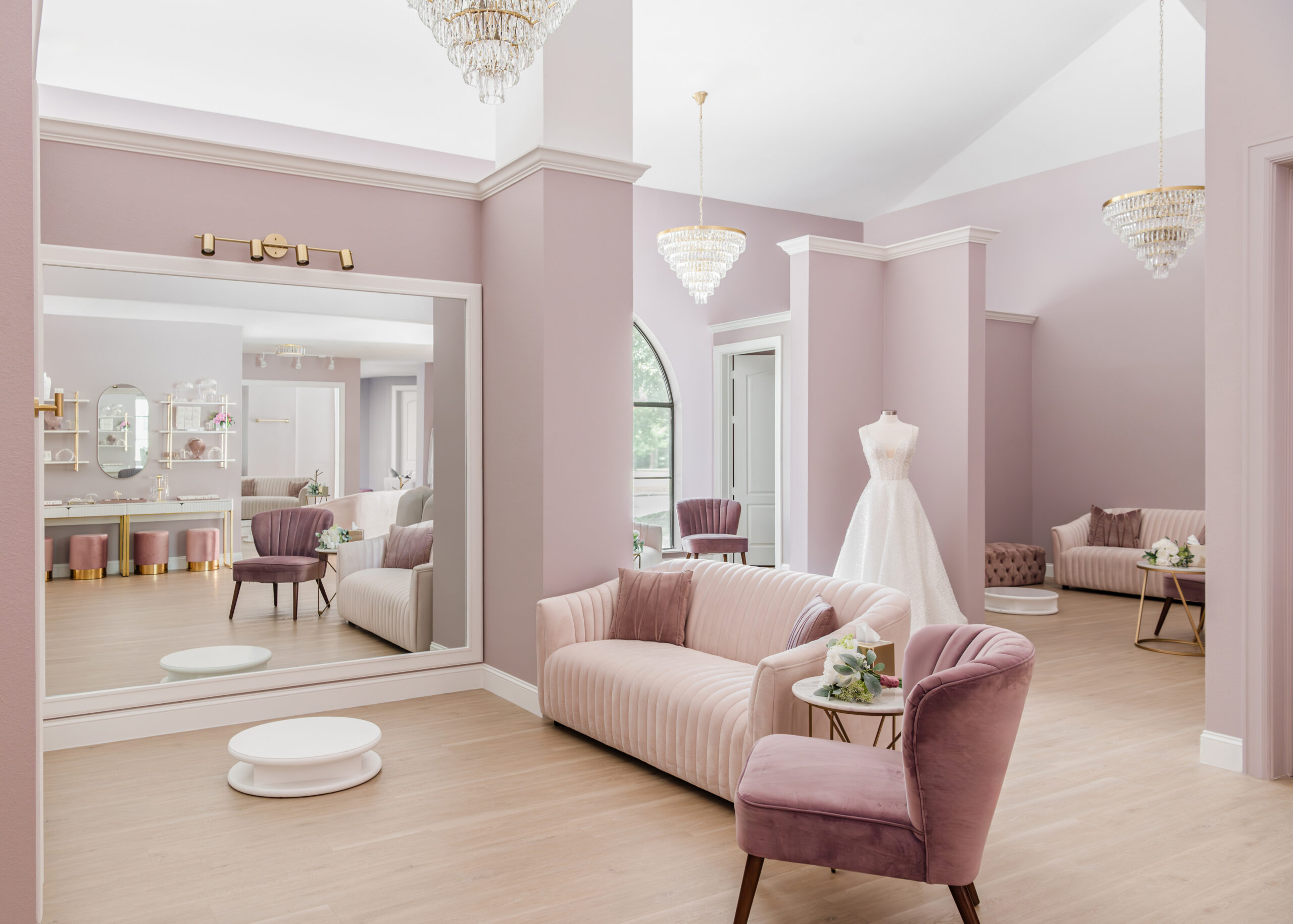 A bridal boutique's open space with soft lavender walls, a crystal chandelier, and comfortable seating areas. A mannequin dressed in a wedding gown stands to the right, with ample mirrors reflecting the airy environment.