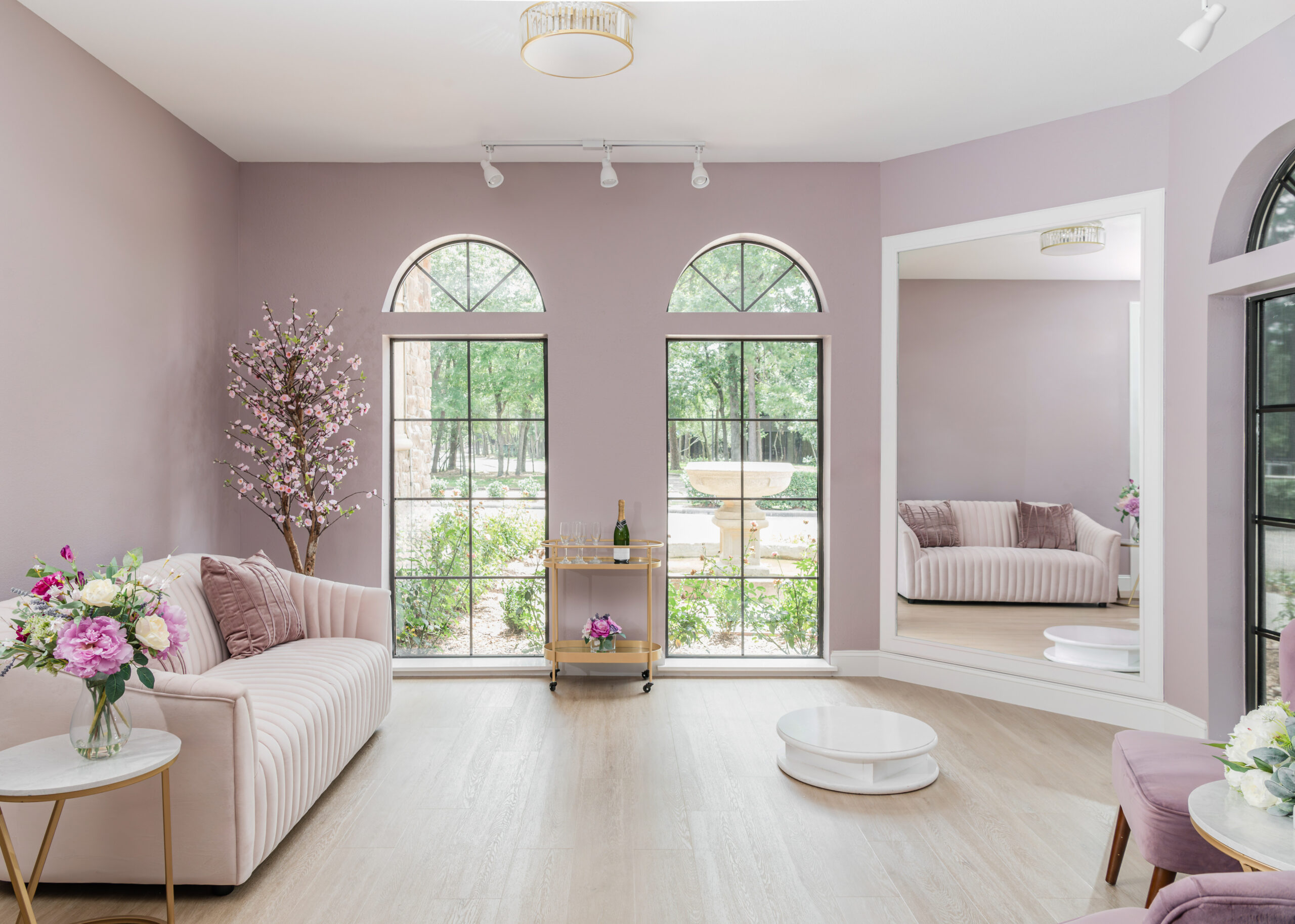 An elegant waiting area in a bridal shop featuring two arched windows with greenery outside. The room includes a blush pink sofa, a flower arrangement on a small table, and a golden bar cart with champagne glasses.