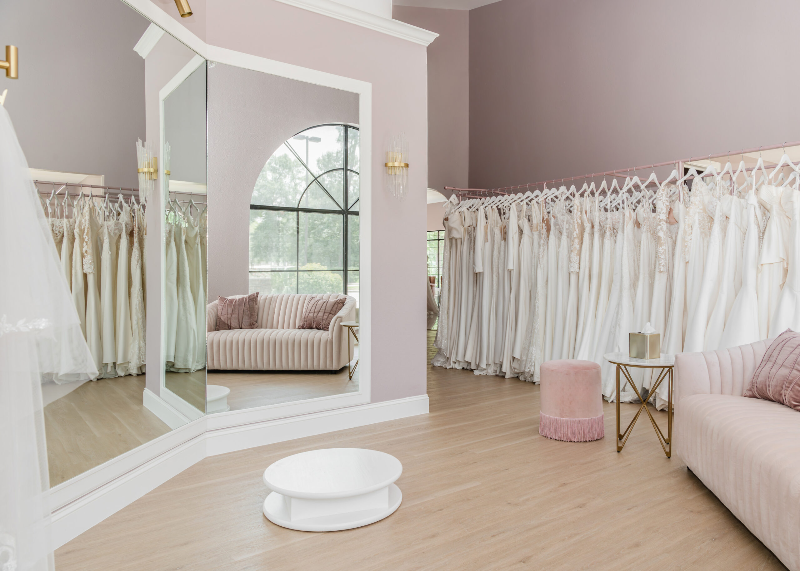 A well-lit bridal fitting room with a large, arched mirror and a soft pink sofa. Rows of wedding gowns are neatly displayed on racks against a pastel wall, creating a serene and welcoming atmosphere.