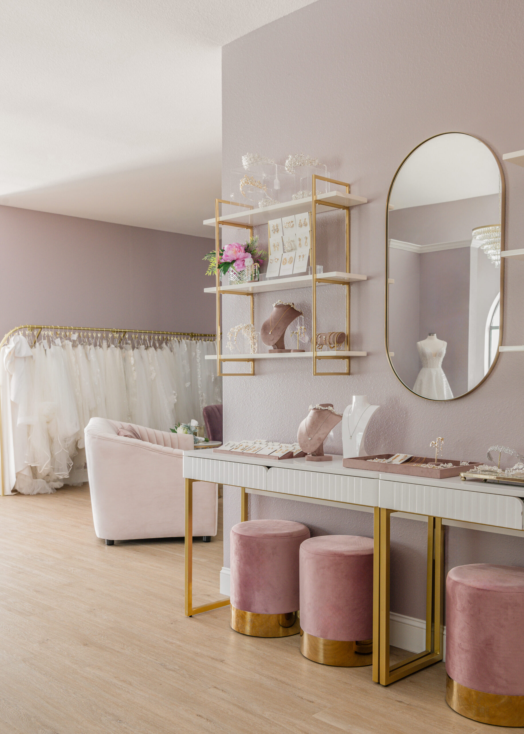 A stylish display area in a bridal boutique featuring a jewelry counter with a mirror, pink stools, and shelving with bridal accessories against a light purple wall.