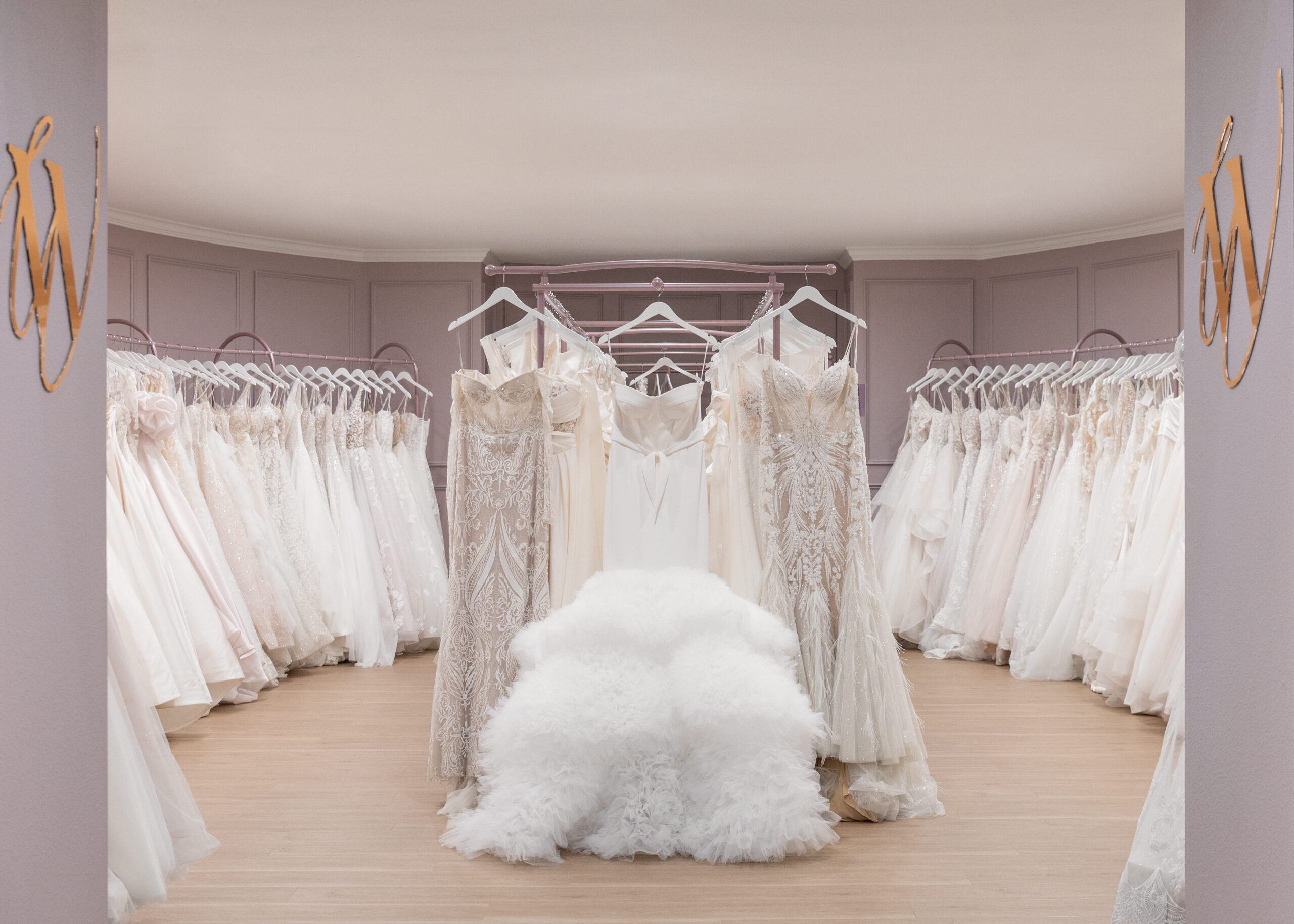 A spacious bridal boutique showcasing a variety of white and ivory wedding dresses hanging on racks, arranged in a semi-circular fashion. In the center, a fluffy white tulle chair adds an elegant touch to the room.