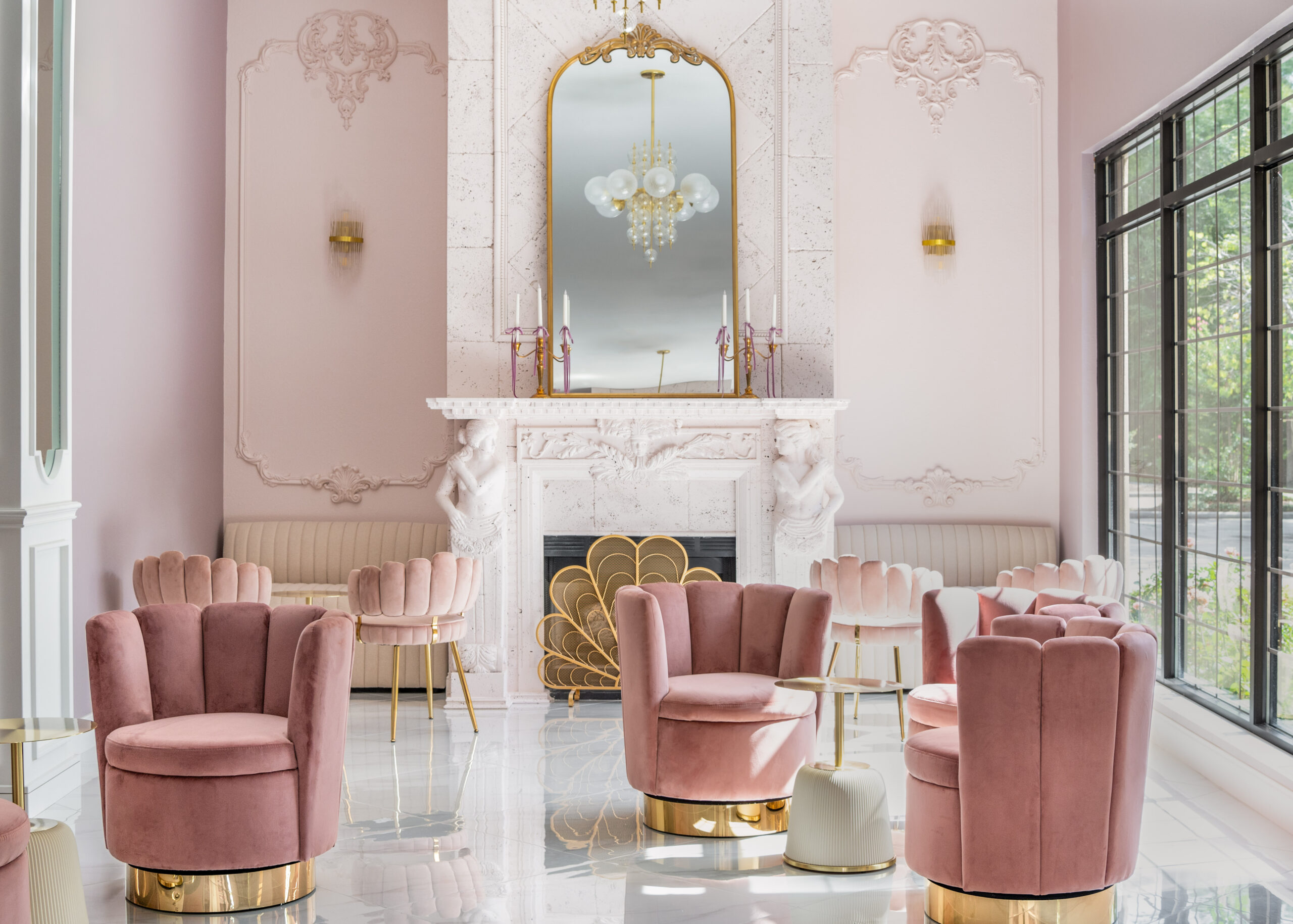 A spacious seating area in a bridal boutique with pink velvet chairs, a marble fireplace with gold accents, and chandeliers.