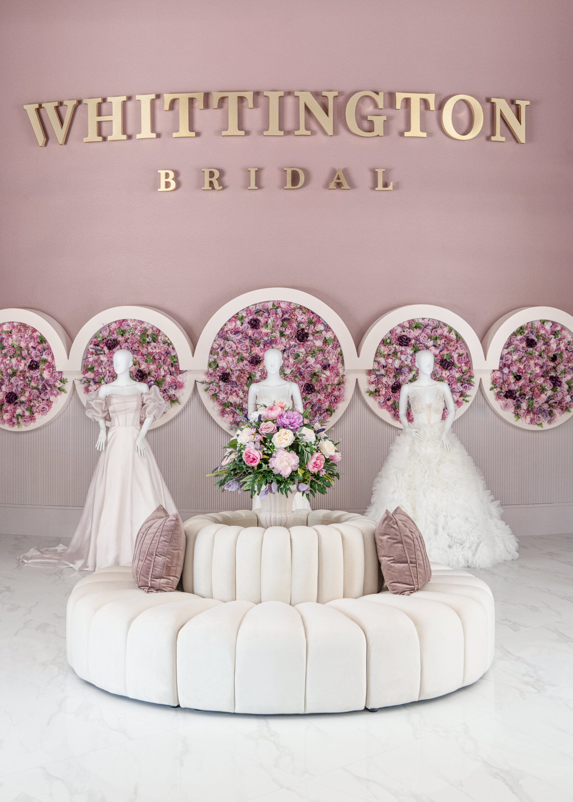 The interior of a bridal boutique featuring mannequins in wedding dresses with a floral backdrop, and a circular white couch beneath a sign that reads "Whittington Bridal."