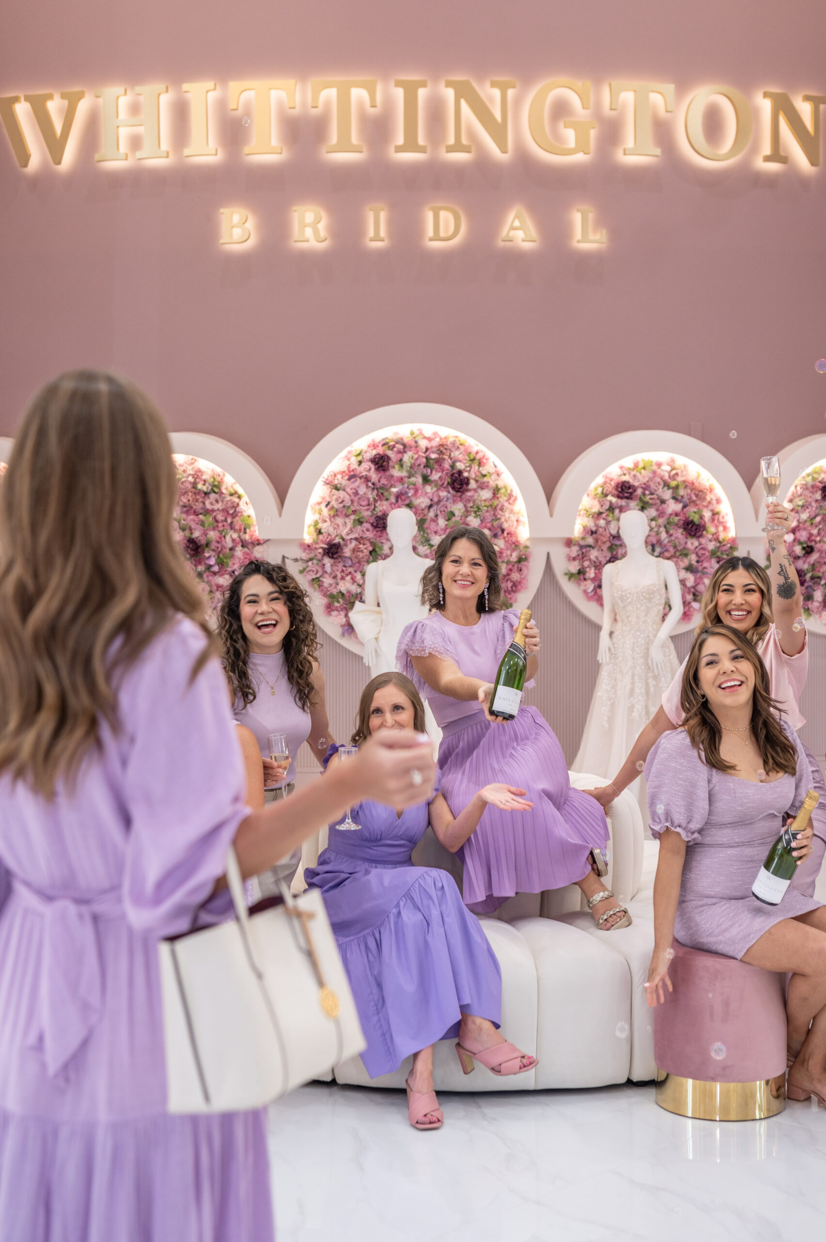 A group of women seated on cushioned stools in a bridal boutique, holding champagne bottles and glasses, smiling and celebrating under a sign that reads "Whittington Bridal."