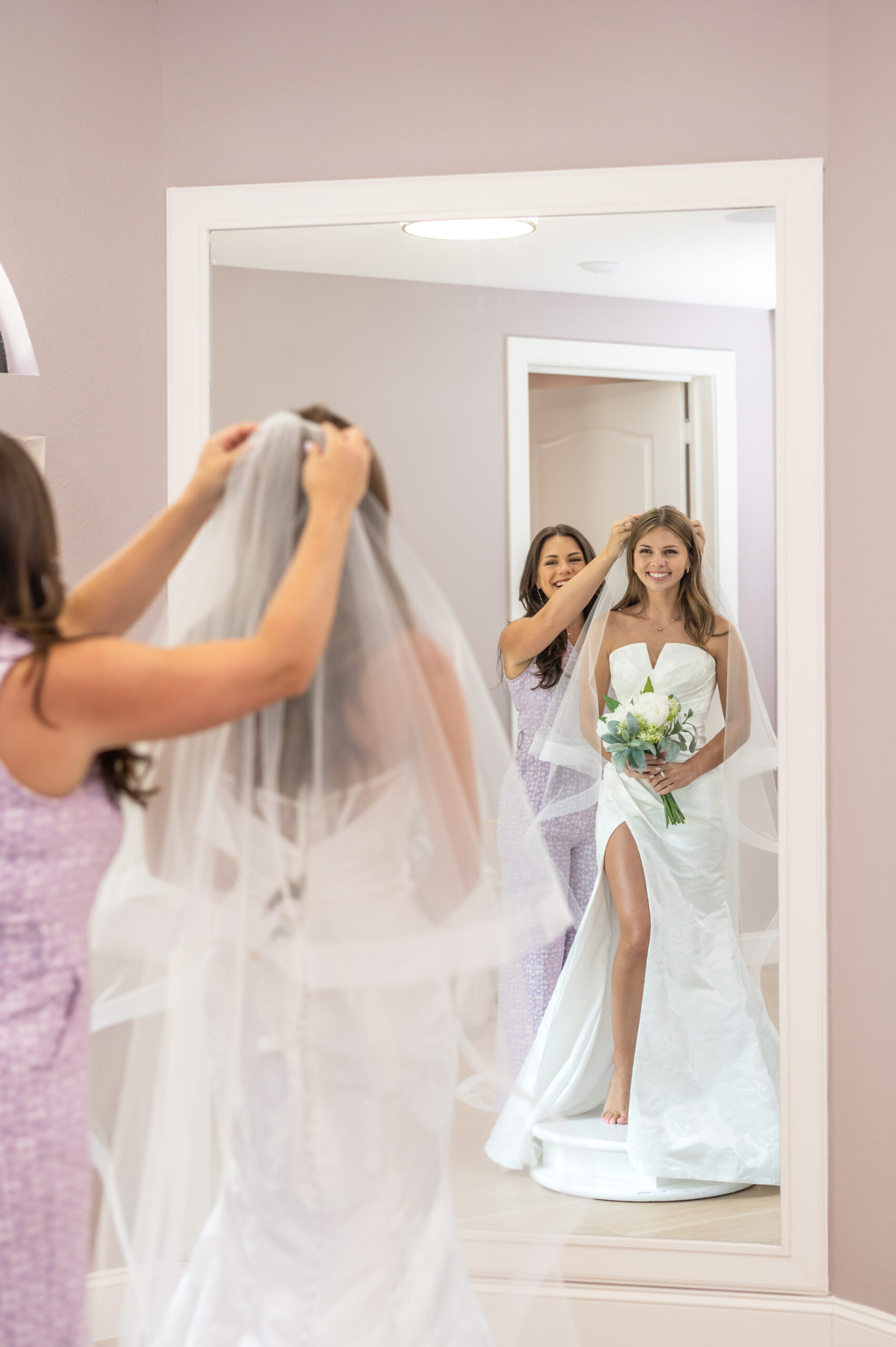 A woman adjusting the veil of a bride-to-be who is holding a bouquet and looking in a mirror while standing on a platform in a bridal boutique.