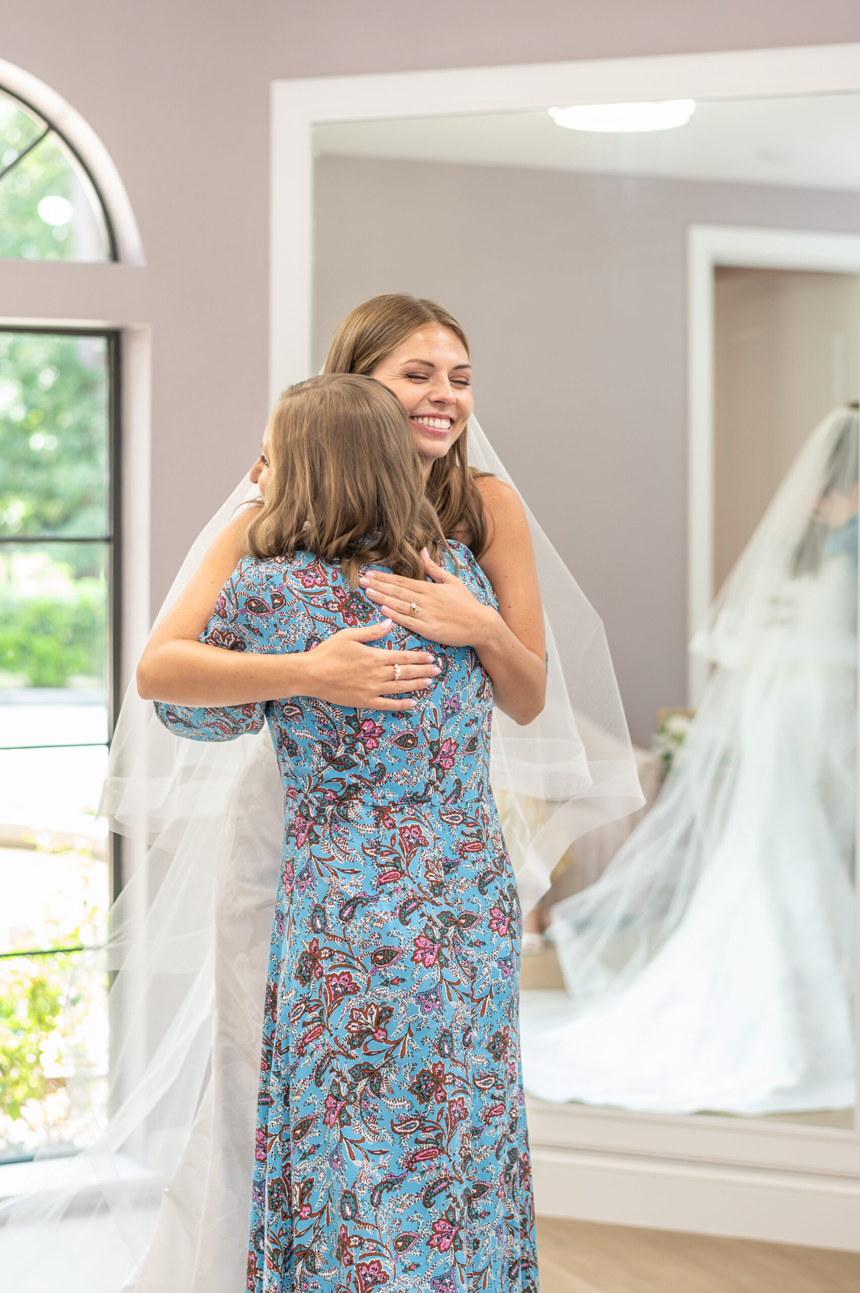 A bride-to-be in a white gown hugging another woman in a floral dress, standing in front of a mirror in a bridal boutique.