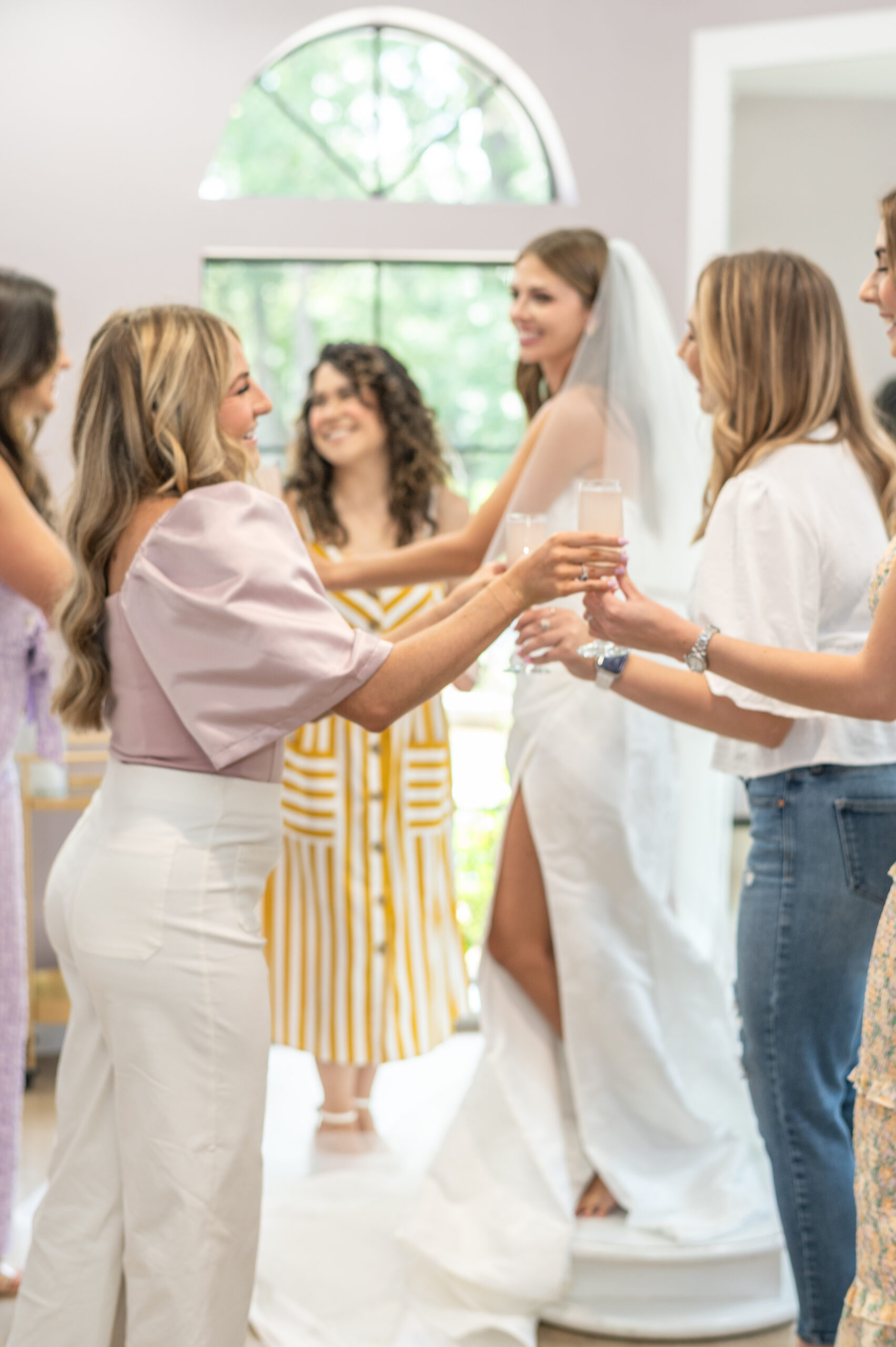 A bride-to-be in a white gown and veil, smiling as she stands on a platform surrounded by friends toasting with champagne glasses in a bridal boutique.