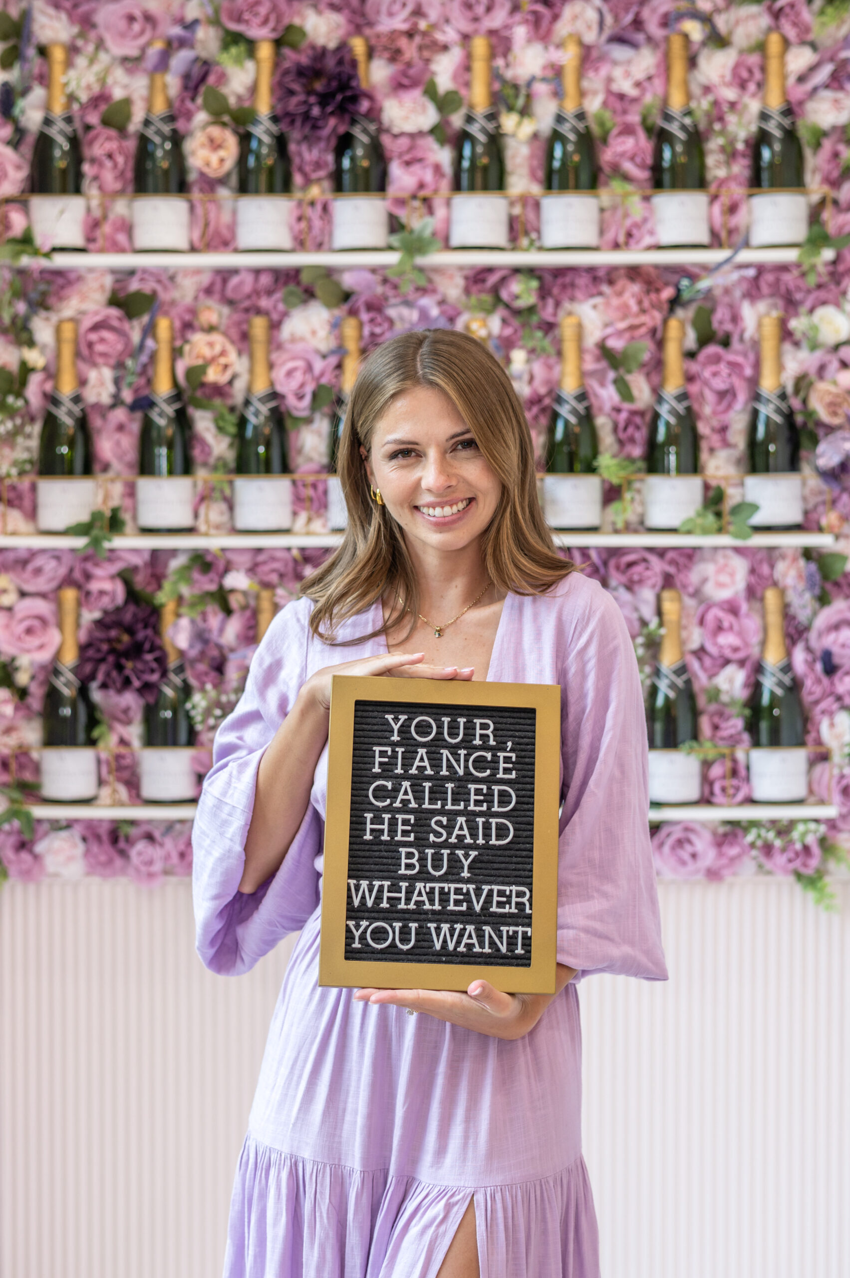 A woman in a lavender dress smiling and holding a sign that says, "Your fiancé called, he said buy whatever you want," with a floral backdrop in the bridal boutique.