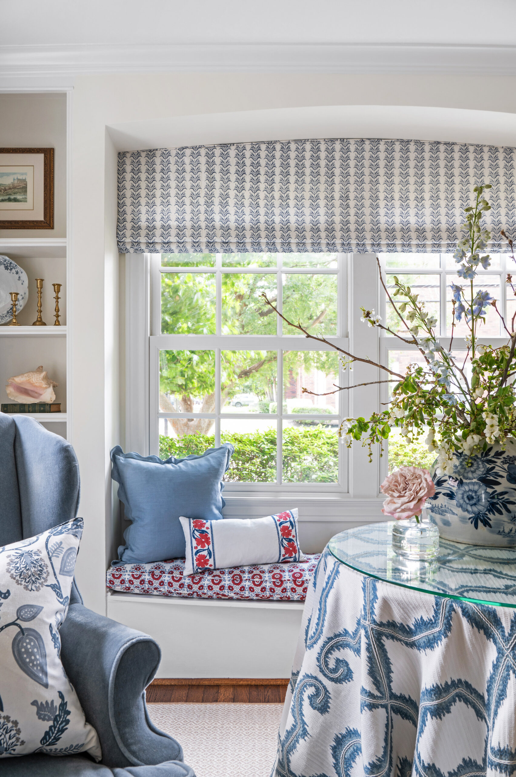 A cozy window nook adorned with vibrant blue and red pillows, accompanied by a round table draped in a beautifully patterned fabric. The room features serene natural light streaming through the windows, enhancing the inviting and charming ambiance. This space would be ideal for reading or relaxing, and is a perfect example of elegant yet comfortable design.