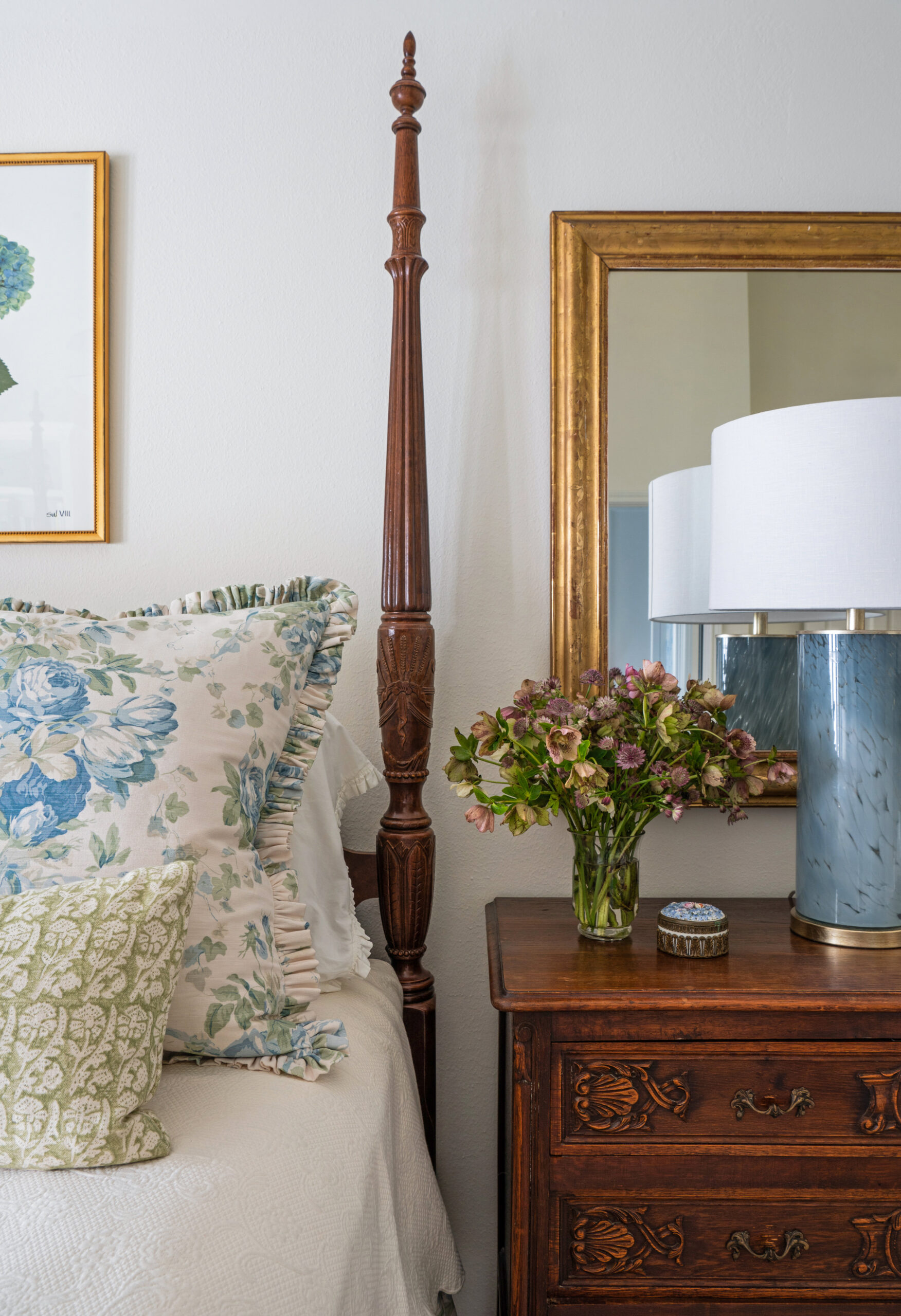 Close-up detail of the bedside table, featuring a dark wood chest with a marble lamp and a delicate floral arrangement. The vintage mirror above adds an element of timelessness to the design. The soft blue, green, and floral patterns evoke a calm, serene atmosphere.