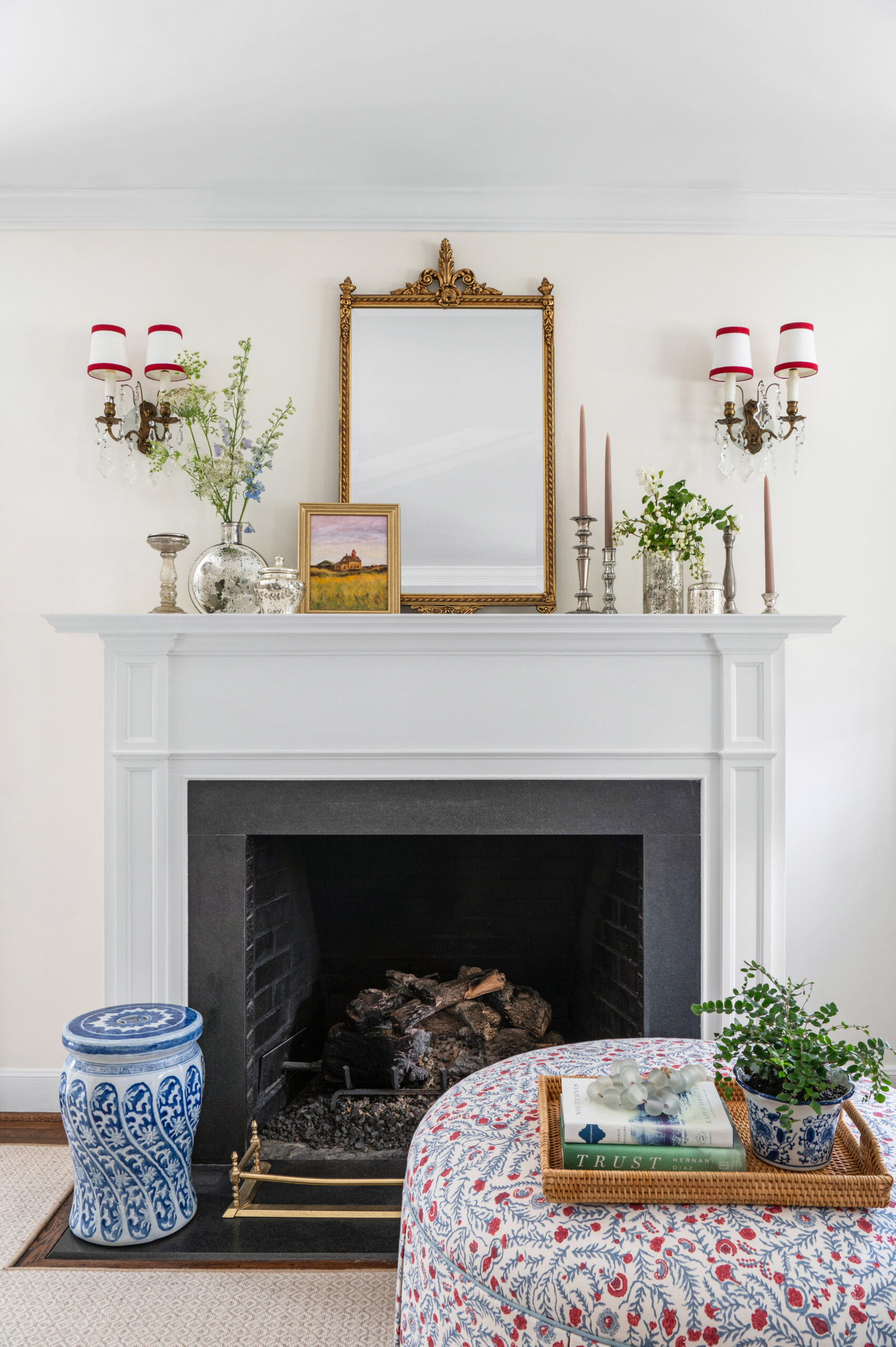 A crisp and clean fireplace area featuring a white mantle and black insert. A mirror above the mantle adds height, while symmetrical accents like sconces and floral arrangements bring balance to the space, enhancing the room's timeless appeal.