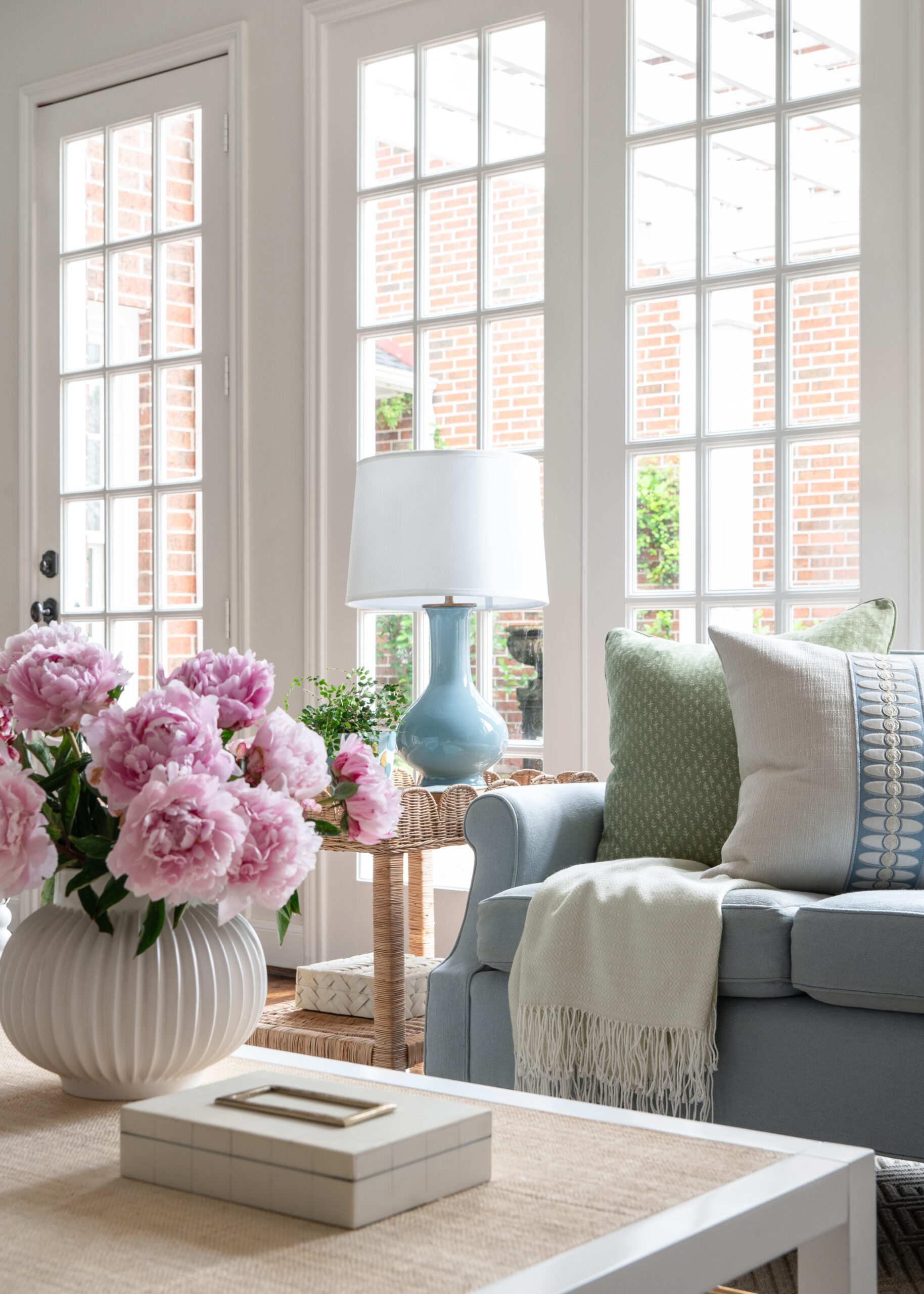 Living room corner with floral accents and natural light: The image focuses on a corner with light streaming in from tall French doors, illuminating a soft blue sofa adorned with pastel throw pillows and a side table with a vase of pink flowers, exuding a sense of tranquility.