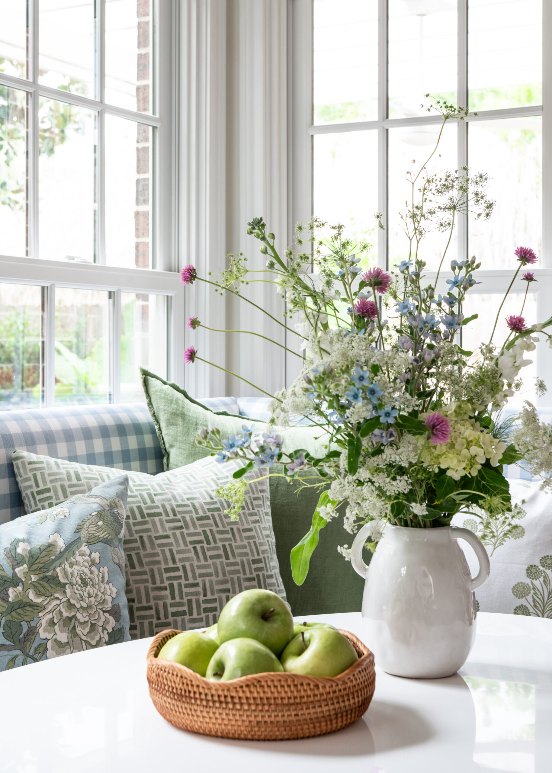 A cozy breakfast nook bathed in natural light, featuring a gingham-patterned bench with green and blue pillows. A vase filled with wildflowers adds charm, while a wicker bowl with fresh green apples completes the inviting atmosphere. Interior Design Photography Licensing allows these beautifully styled spaces to be shared and enjoyed by a wider audience.