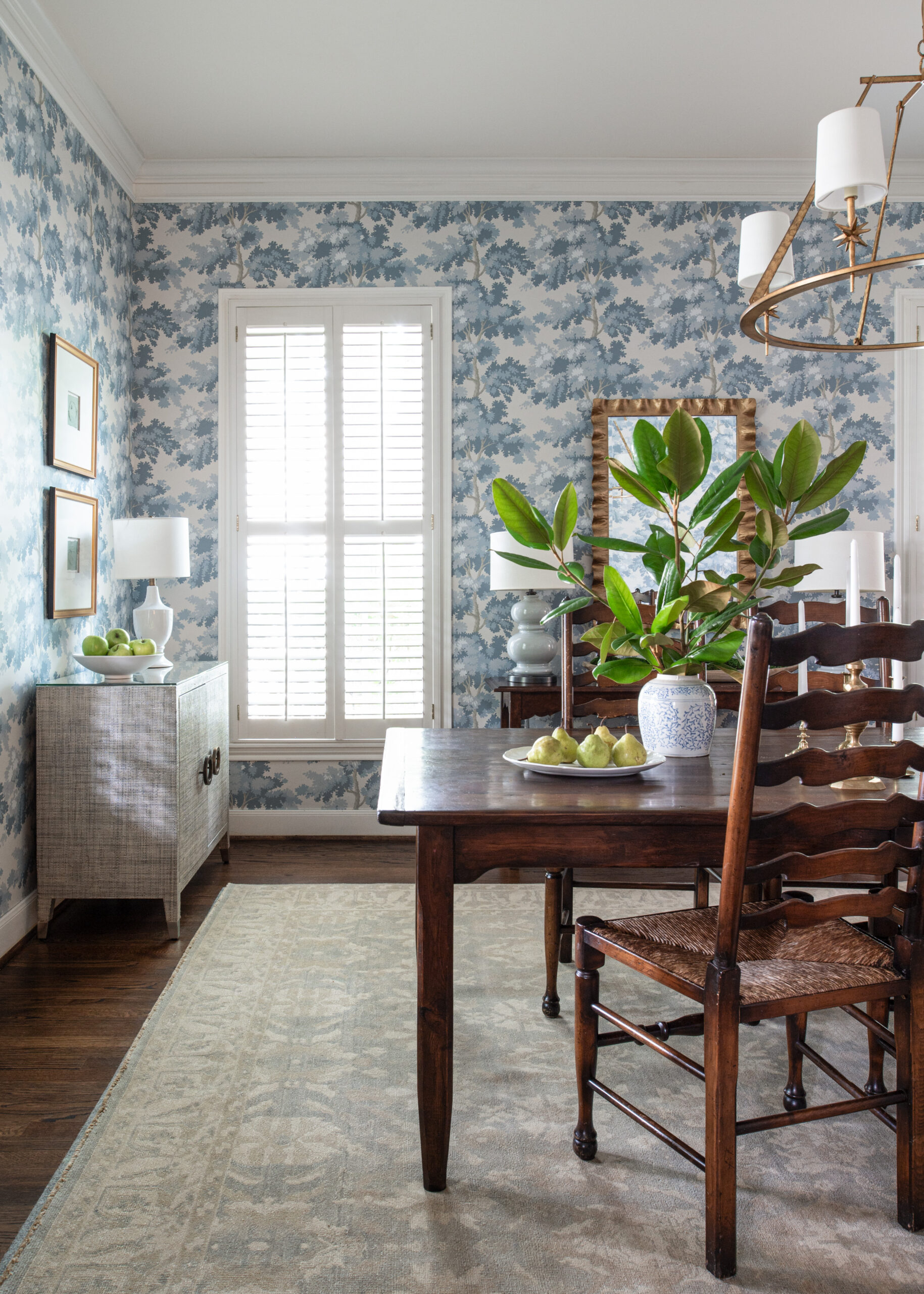 A sophisticated dining room with soft blue and white wallpaper, adorned with rustic wooden furniture and a brass chandelier. The room is enhanced by the greenery of potted plants and bowls of fresh fruit, creating a perfect blend of elegance and warmth.