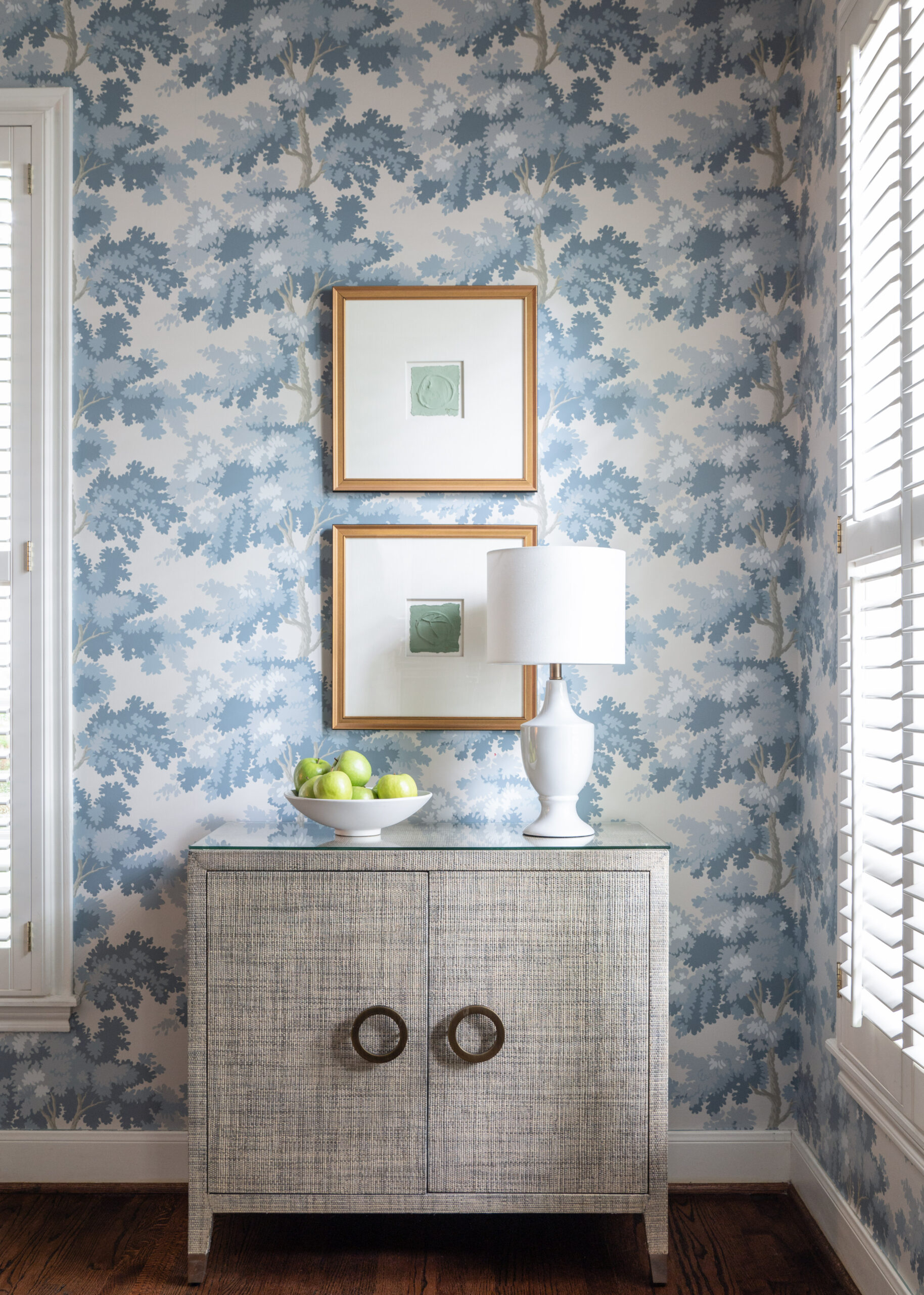 Close-up of a dining room corner featuring a light grey textured console topped with a white lamp and a bowl of green apples. The floral wallpaper in blue adds a soft touch to the room, offering a balance of modernity and tradition. This space is ideal for showcasing Interior Design Photography Licensing, where every detail speaks to the artistry of home decor.