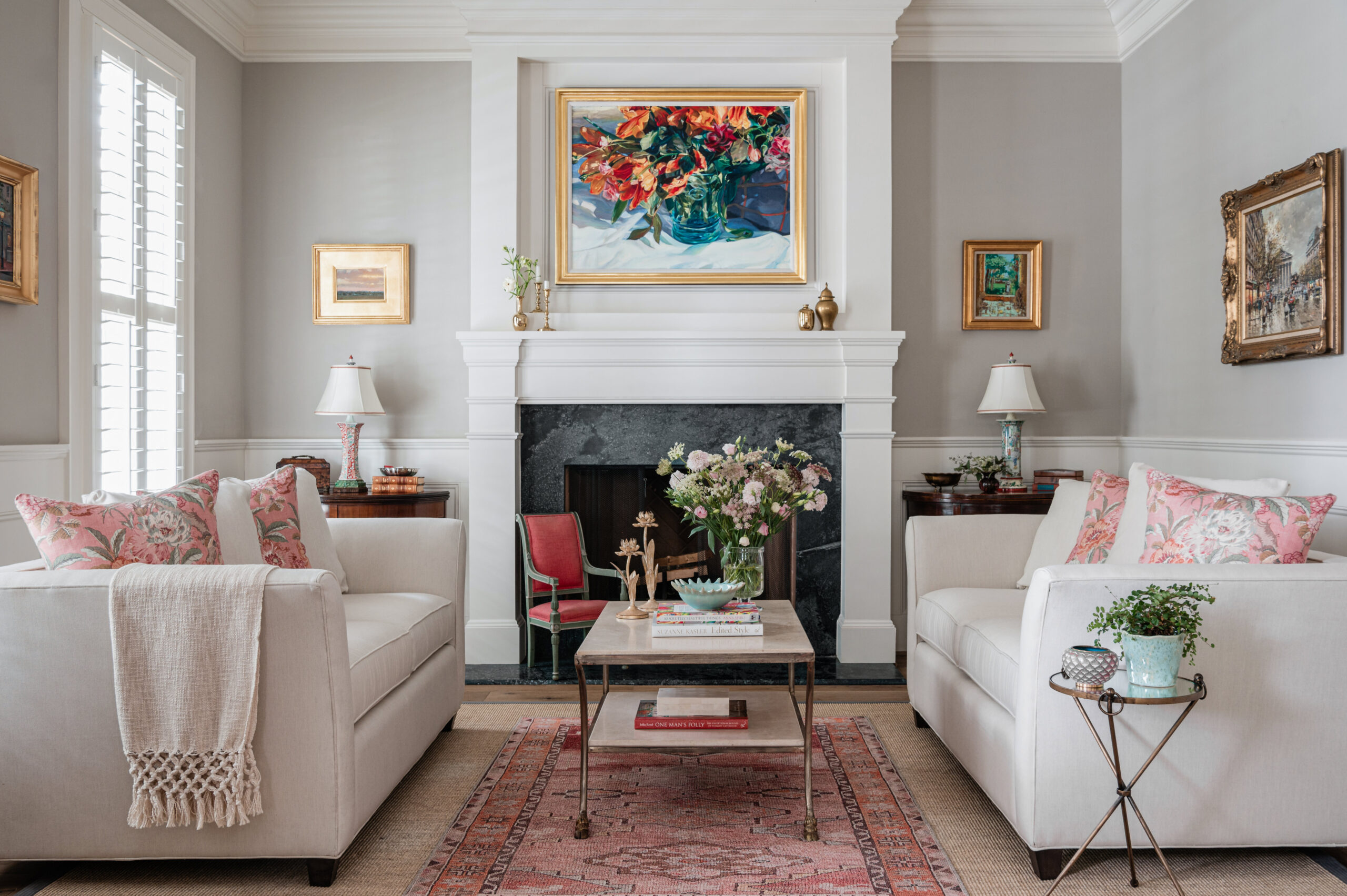 A cozy living room with a soft white couch adorned with floral patterned pillows. A marble coffee table features a vase filled with fresh pink and white flowers. The room is tastefully decorated with paintings, including a vibrant floral painting above the mantel. The warm, elegant setting highlights Interior Design Photography Licensing, capturing the beauty and attention to detail of the room's decor.