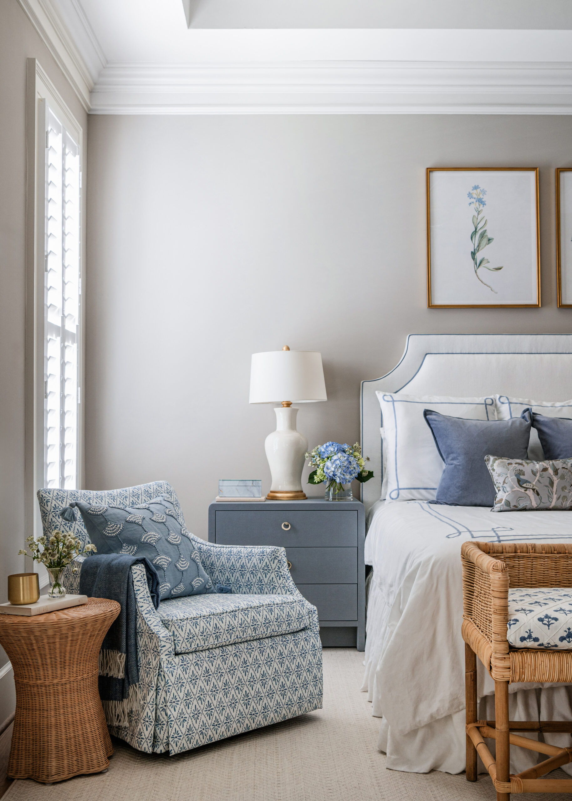 A serene bedroom featuring soft blue tones and classic design elements: This image showcases a cozy bedroom setup with a blue upholstered bed, an armchair in matching patterned fabric, and a wicker side table. The light streaming through the window highlights the elegant design, making it perfect for showcasing Interior Design Photography Licensing.