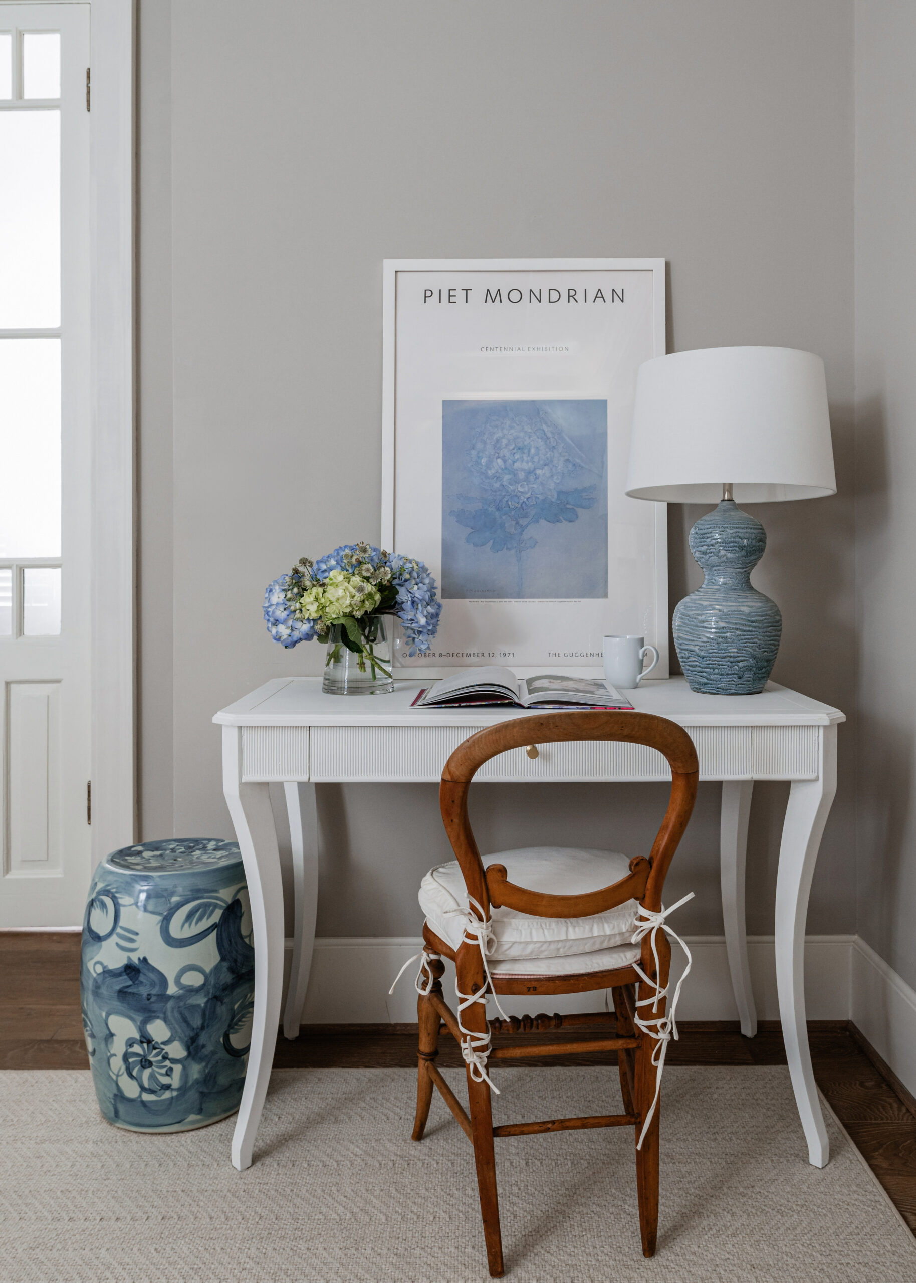 Bright and minimalist home office corner with white furniture: A small white desk is styled with a chair and soft blue accents like a table lamp and hydrangeas, complemented by a framed Piet Mondrian print, making this space feel cozy and organized. The simple yet artistic layout of this interior is ideal for Interior Design Photography Licensing.