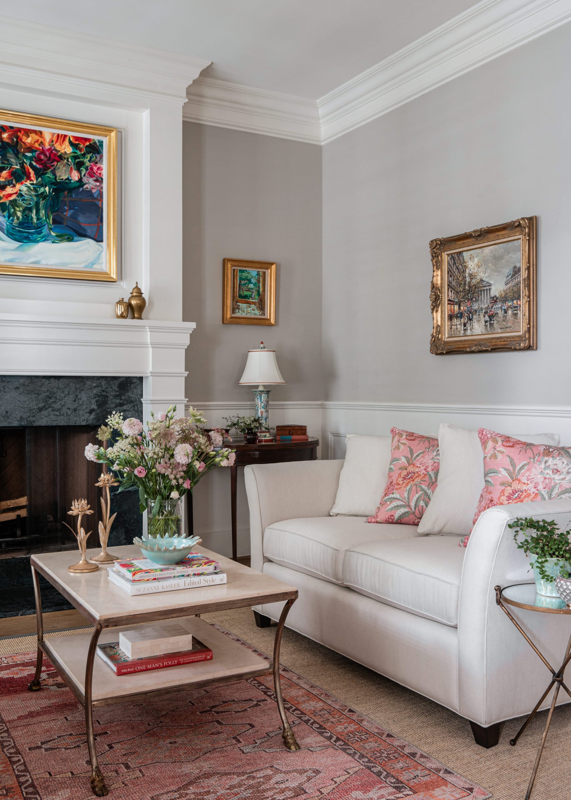 A cozy living room with a soft white couch adorned with floral patterned pillows. A marble coffee table features a vase filled with fresh pink and white flowers. The room is tastefully decorated with paintings, including a vibrant floral painting above the mantel. The warm, elegant setting highlights Interior Design Photography Licensing, capturing the beauty and attention to detail of the room's decor.