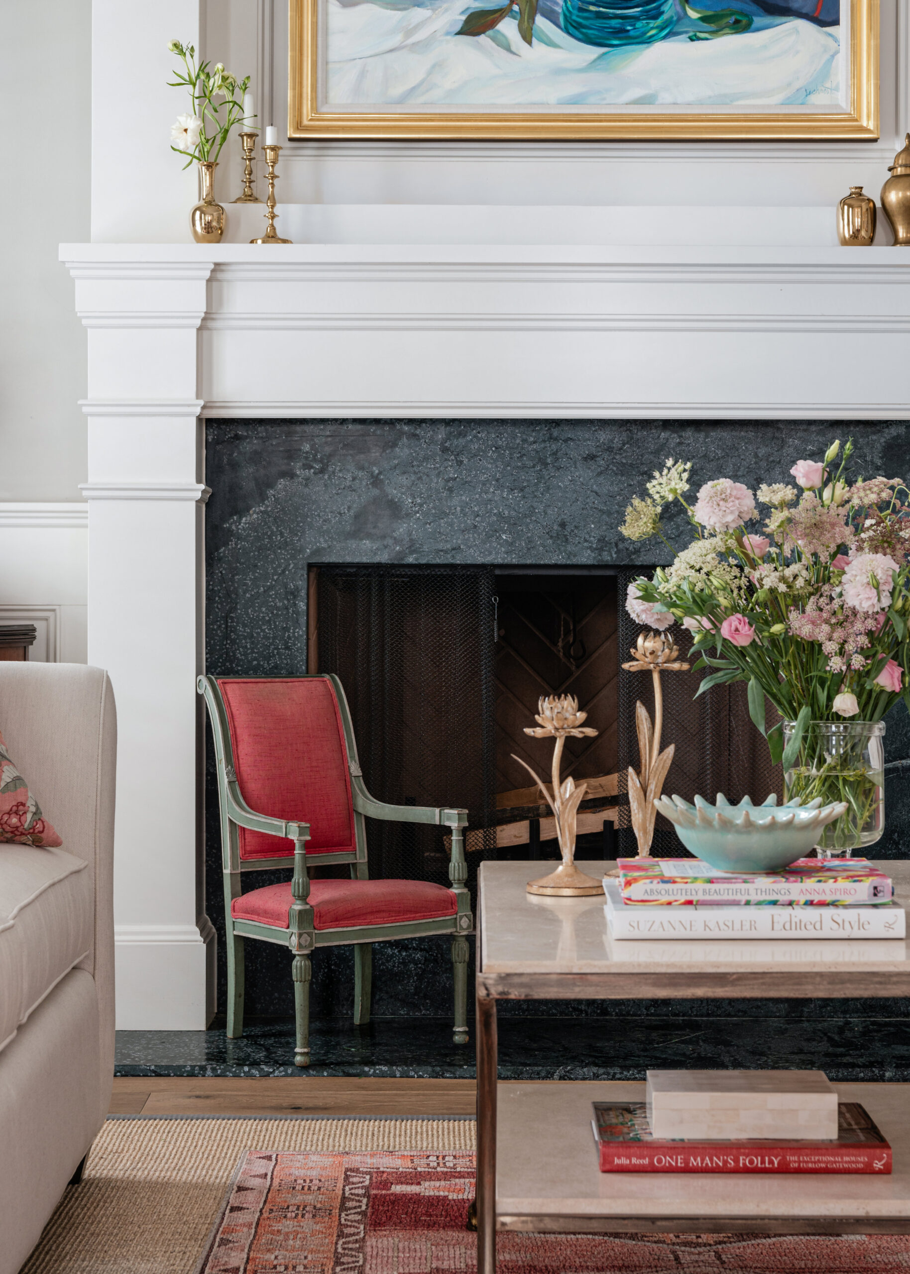 A close-up view of a white fireplace with intricate moldings. A small vintage chair with a red cushion sits to the side, and a beautifully arranged floral vase rests on the marble coffee table. Above the mantel, a colorful framed artwork adds a pop of color to the neutral tones of the room, ideal for Interior Design Photography Licensing purposes.