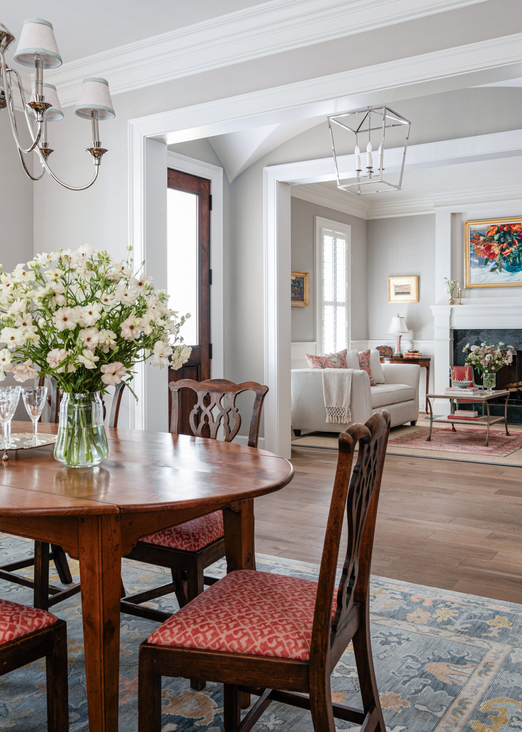 A wide shot of a dining room leading into the living area, featuring a round wooden table with red cushioned chairs. A floral arrangement in the center adds a fresh and elegant touch. The adjacent room showcases a comfortable seating area and a fireplace, providing a seamless flow between spaces. The natural light streaming in enhances the warmth of this classic design.