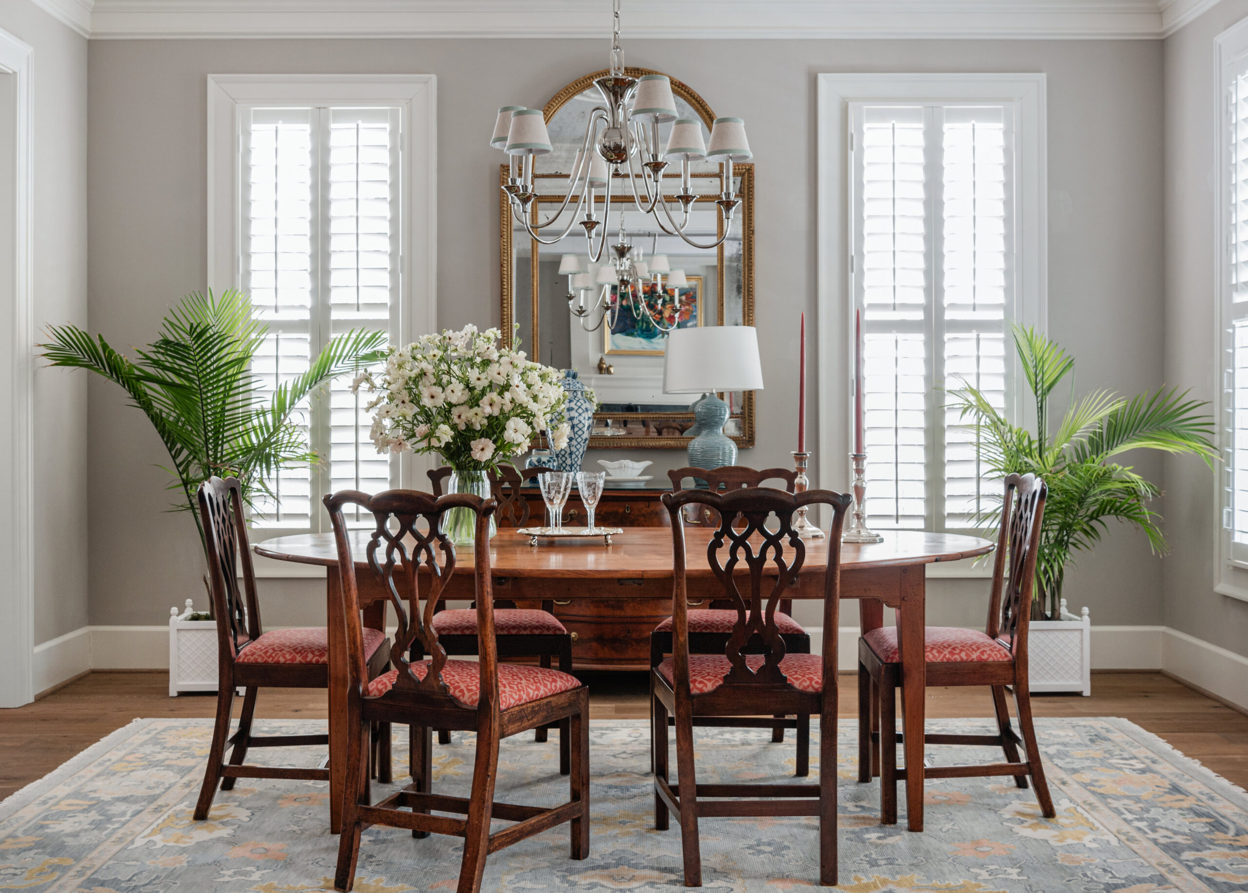 A wide shot of a dining room leading into the living area, featuring a round wooden table with red cushioned chairs. A floral arrangement in the center adds a fresh and elegant touch. The adjacent room showcases a comfortable seating area and a fireplace, providing a seamless flow between spaces. The natural light streaming in enhances the warmth of this classic design