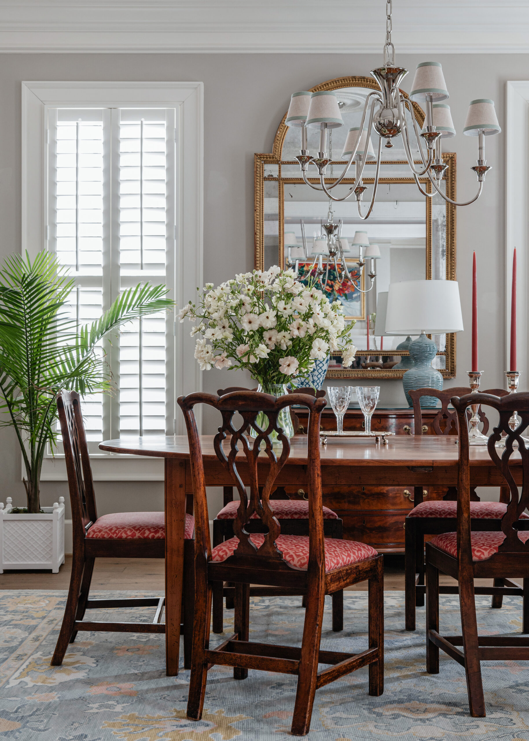 A formal dining room with an antique wooden table and chairs, a large vase of white flowers as the centerpiece, and two symmetrical palm plants adding greenery to the space. The light from the tall windows and chandelier creates a bright and inviting atmosphere, ideal for showcasing refined Interior Design Photography Licensing.