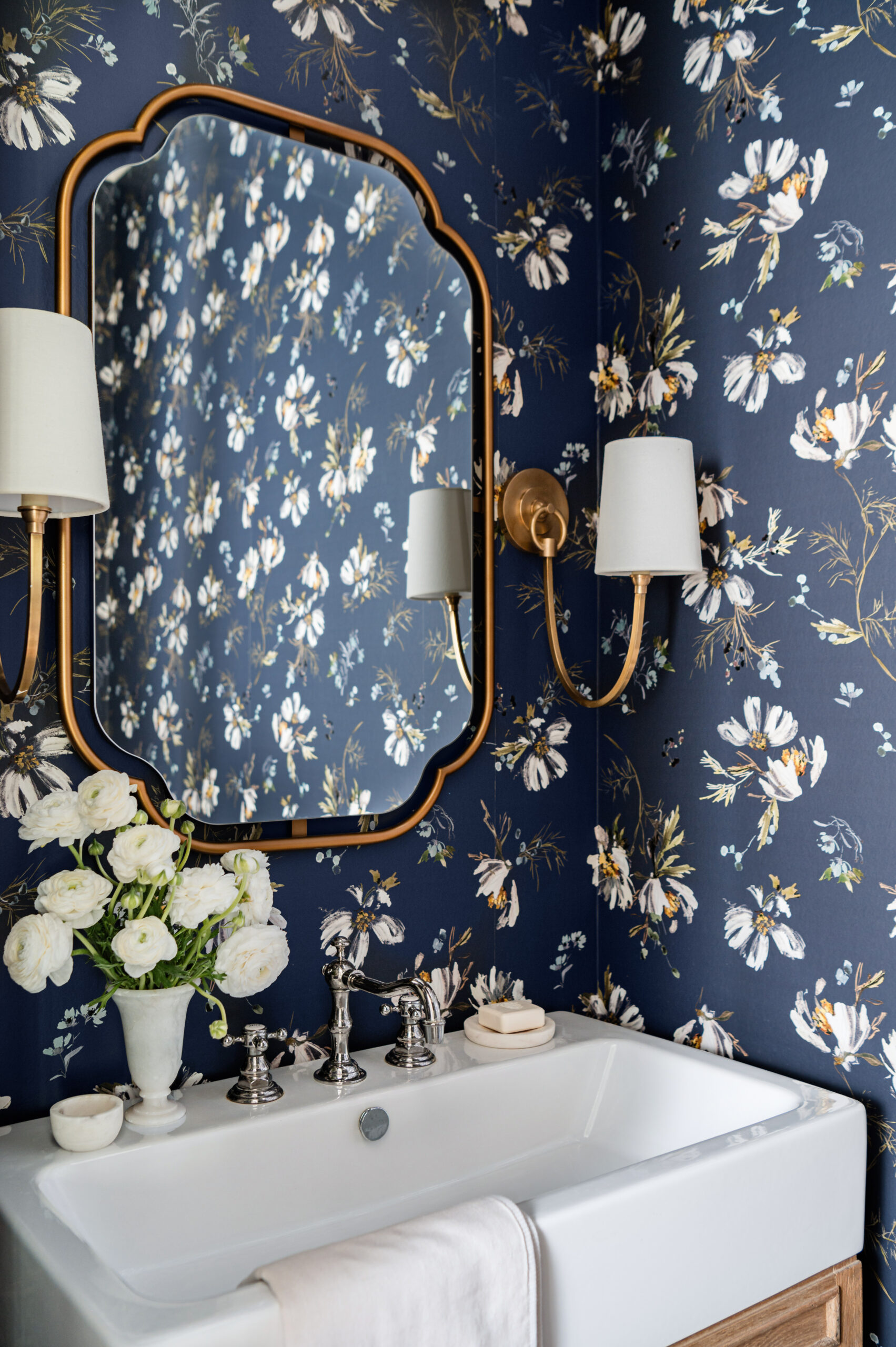 A bold and stylish powder room with a navy floral wallpaper that contrasts beautifully with gold-framed sconces and mirror. The white porcelain sink is paired with polished chrome fixtures, and a bouquet of white flowers adds a soft, elegant touch to this striking design.