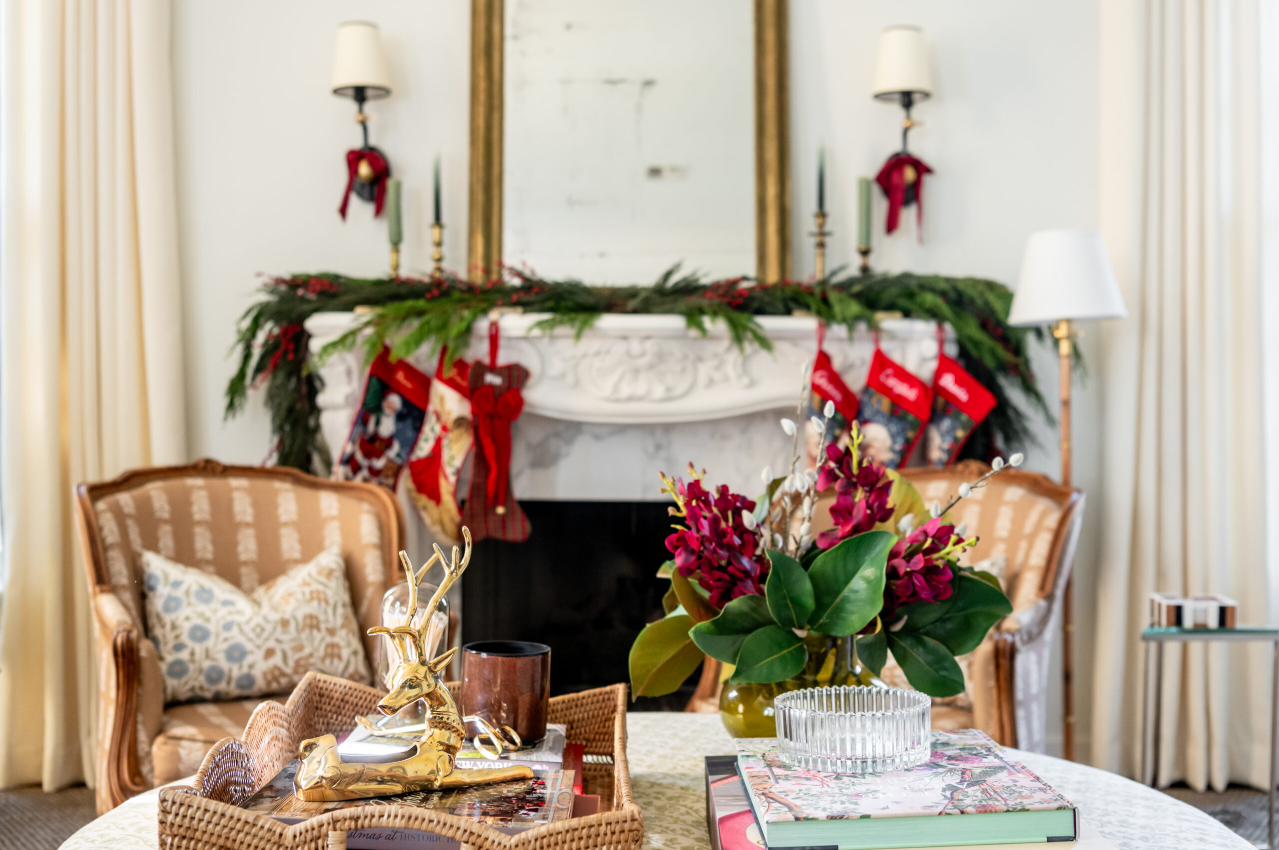 A close-up shot of a tray with seasonal interior design accents, including a golden reindeer, a candle, and holiday books, placed in front of a decorated mantel with stockings