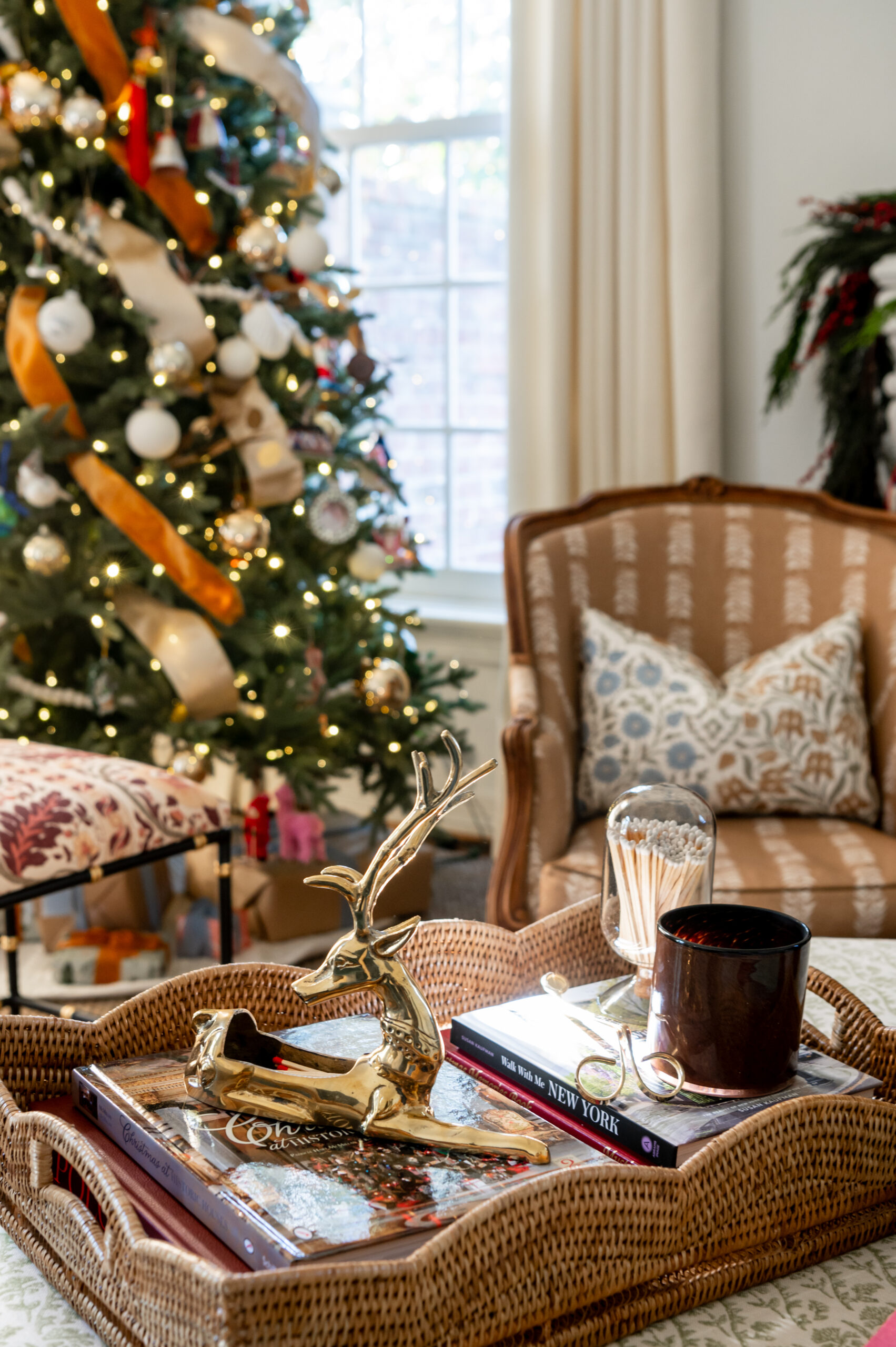 A coffee table with a festive tray showcasing seasonal interior design, including a reindeer decoration, holiday books, and a candle, set against a decorated Christmas tree.