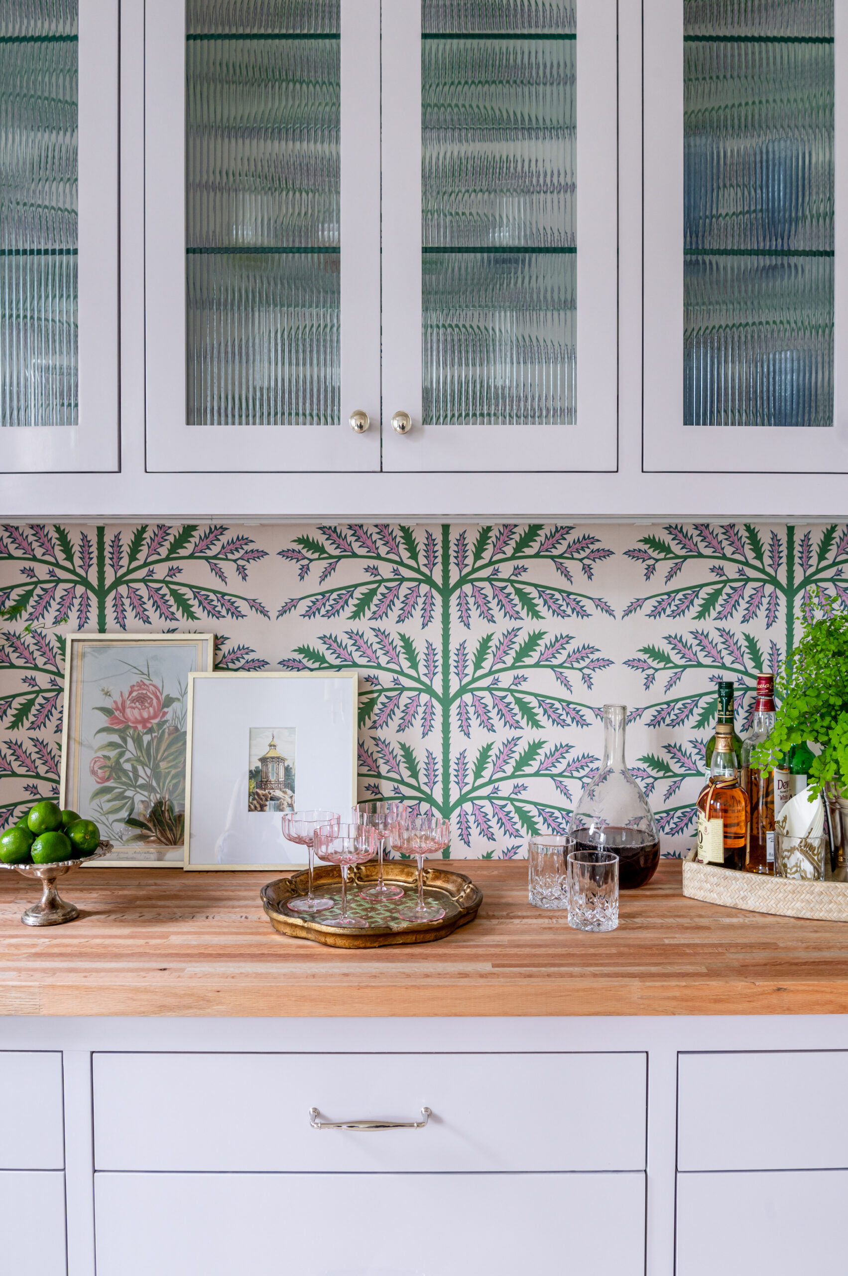 A stylish kitchen with glass-fronted cabinets showcasing bar essentials and vibrant green botanical wallpaper as a backdrop, exuding an inviting atmosphere for entertaining. The master interior design of the space incorporates functional storage with artistic detailing.