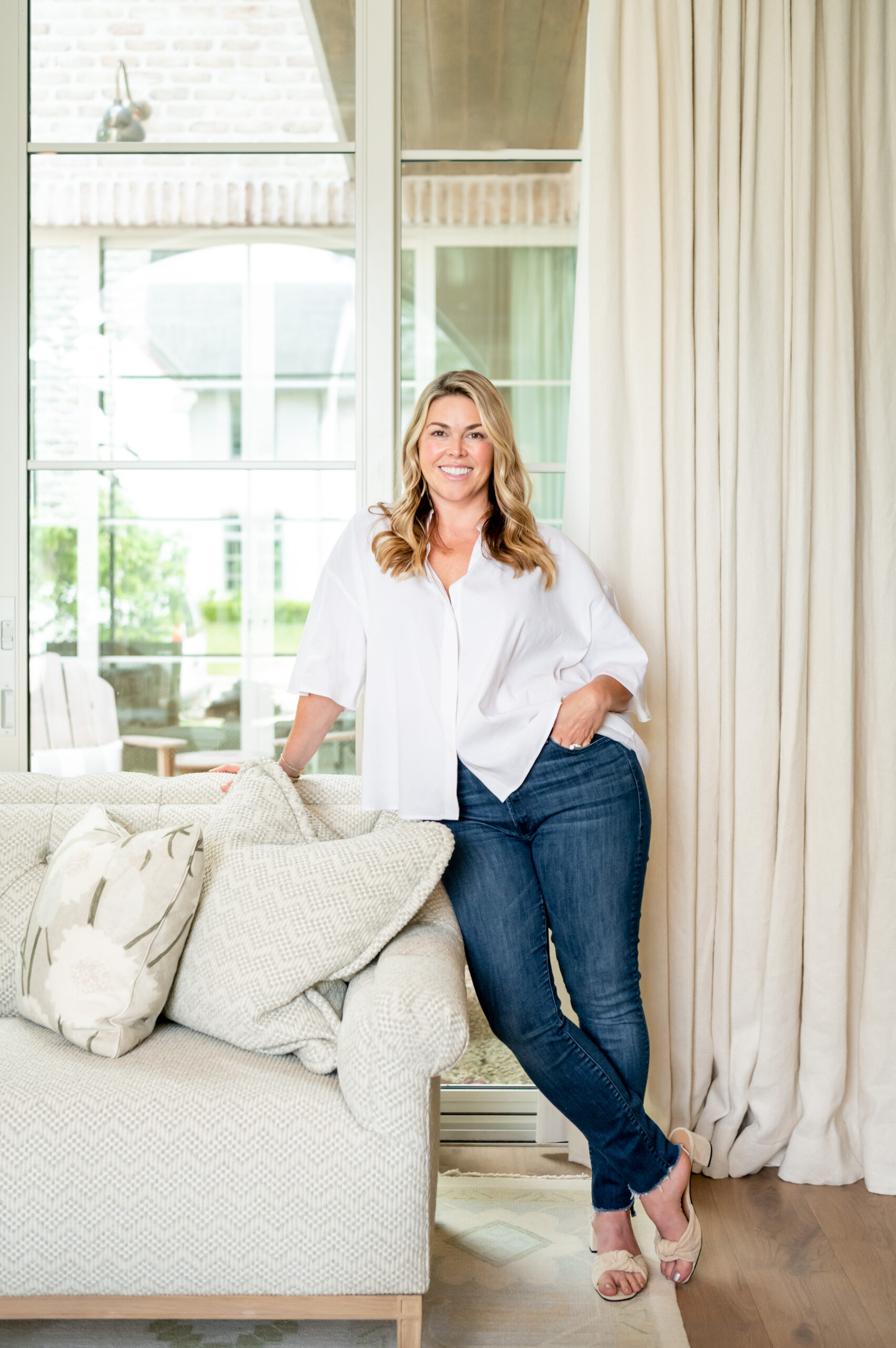 A professional portrait of a woman standing next to a brick fireplace with large windows, showcasing a relaxed yet elegant living space. The room reflects a well-thought-out master interior design with comfortable furnishings and warm tones.