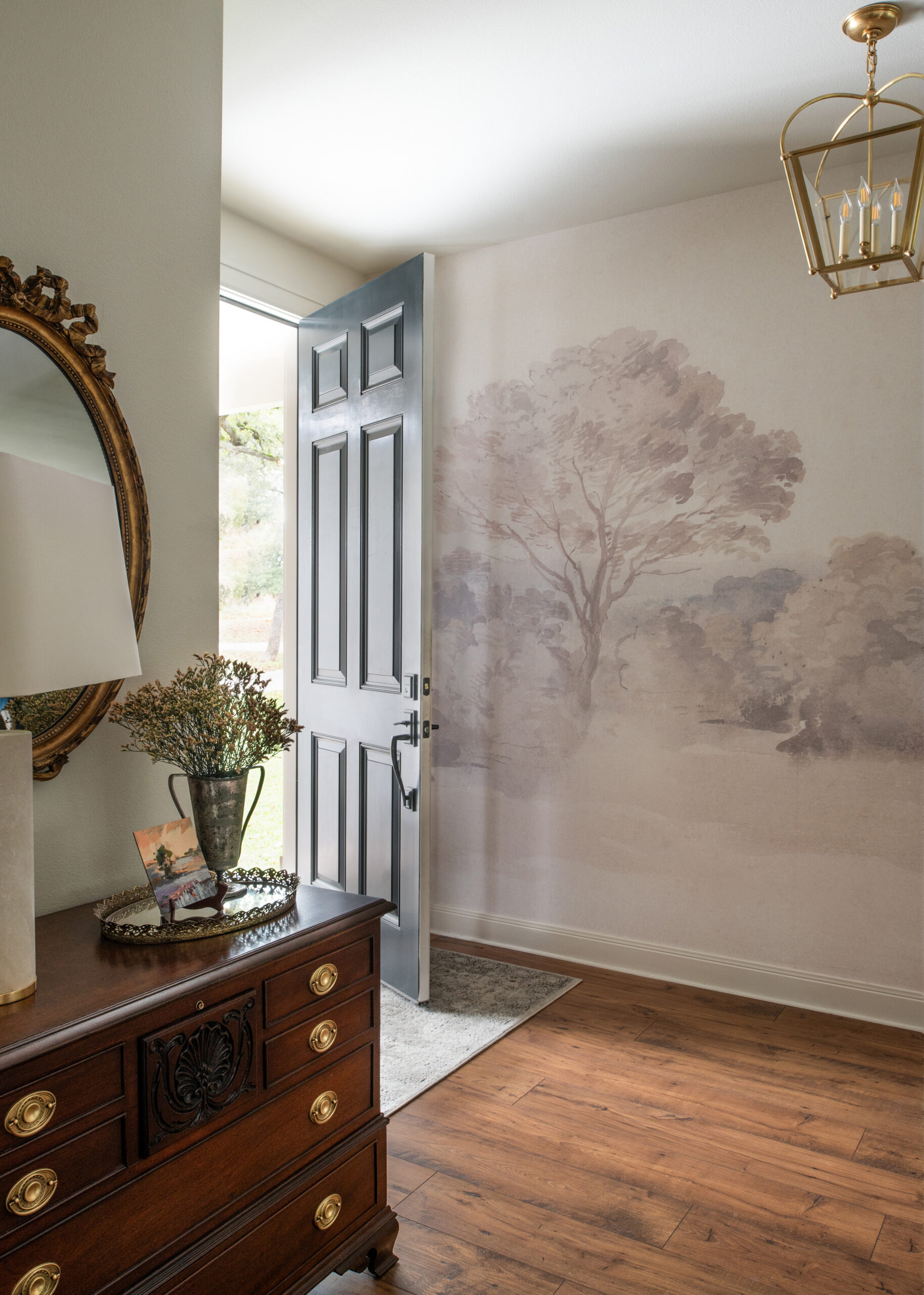 A welcoming entryway with a vintage chest of drawers, neutral-colored walls, and a large door with scenic views. This hallway showcases timeless modern interior design, blending old and new seamlessly.