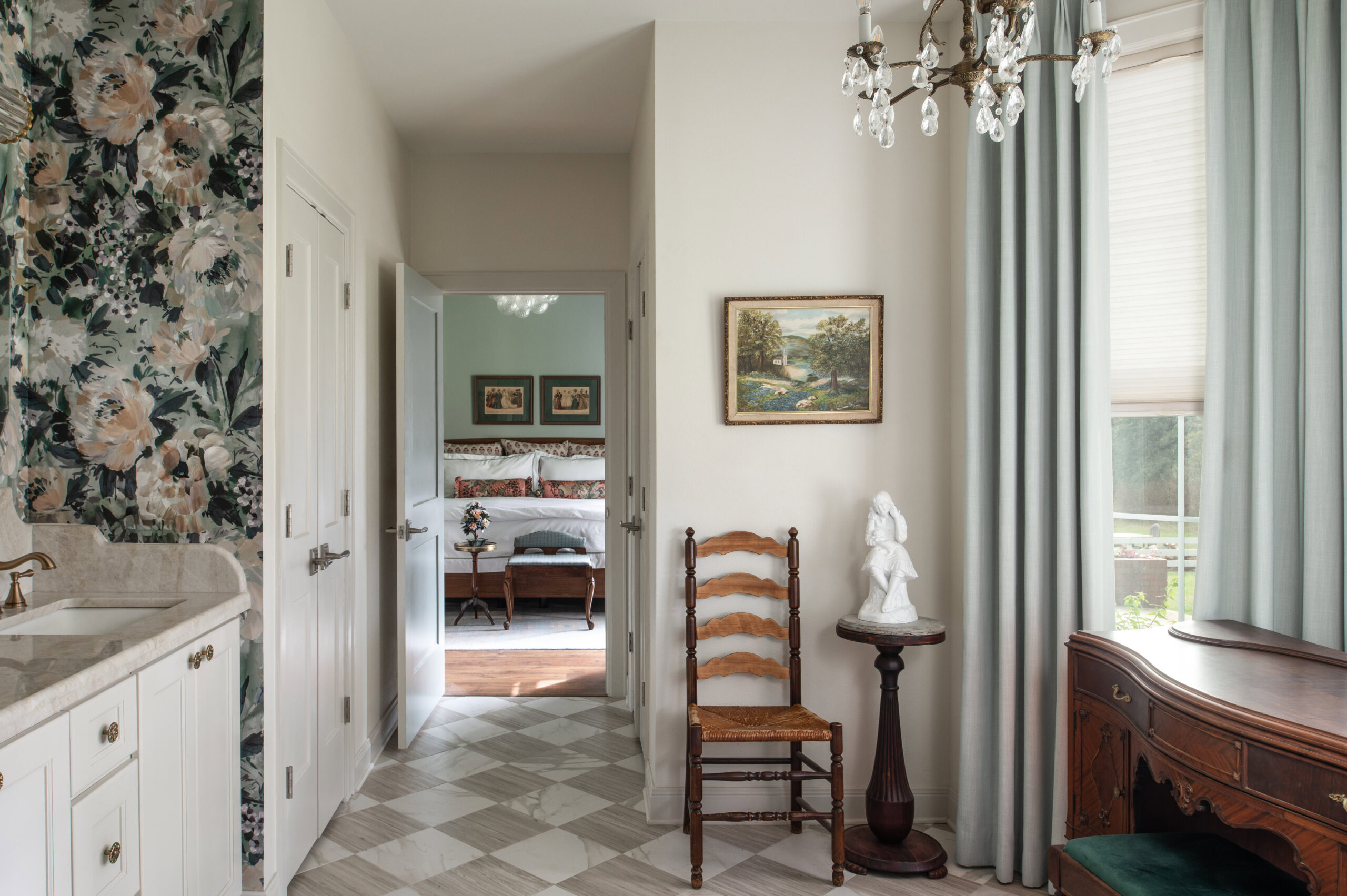 A hallway with a classic chair, artwork, and a soft chandelier that sets a peaceful and timeless modern interior design vibe, with a view into the serene bedroom.