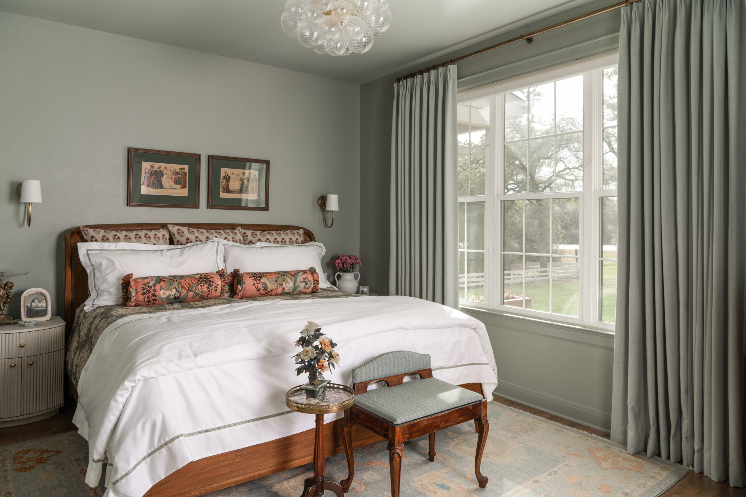 A beautifully designed bedroom with soft green tones, classic art pieces, and a window overlooking the outdoors. The space feels timeless and inviting, blending traditional elegance with modern comfort.