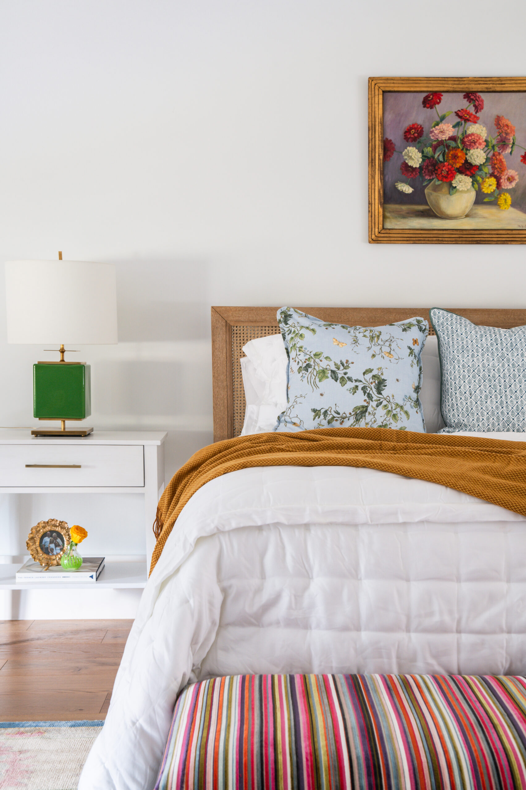 A cozy bedroom with a striped bench, warm blankets, and a colorful painting above the bed.
