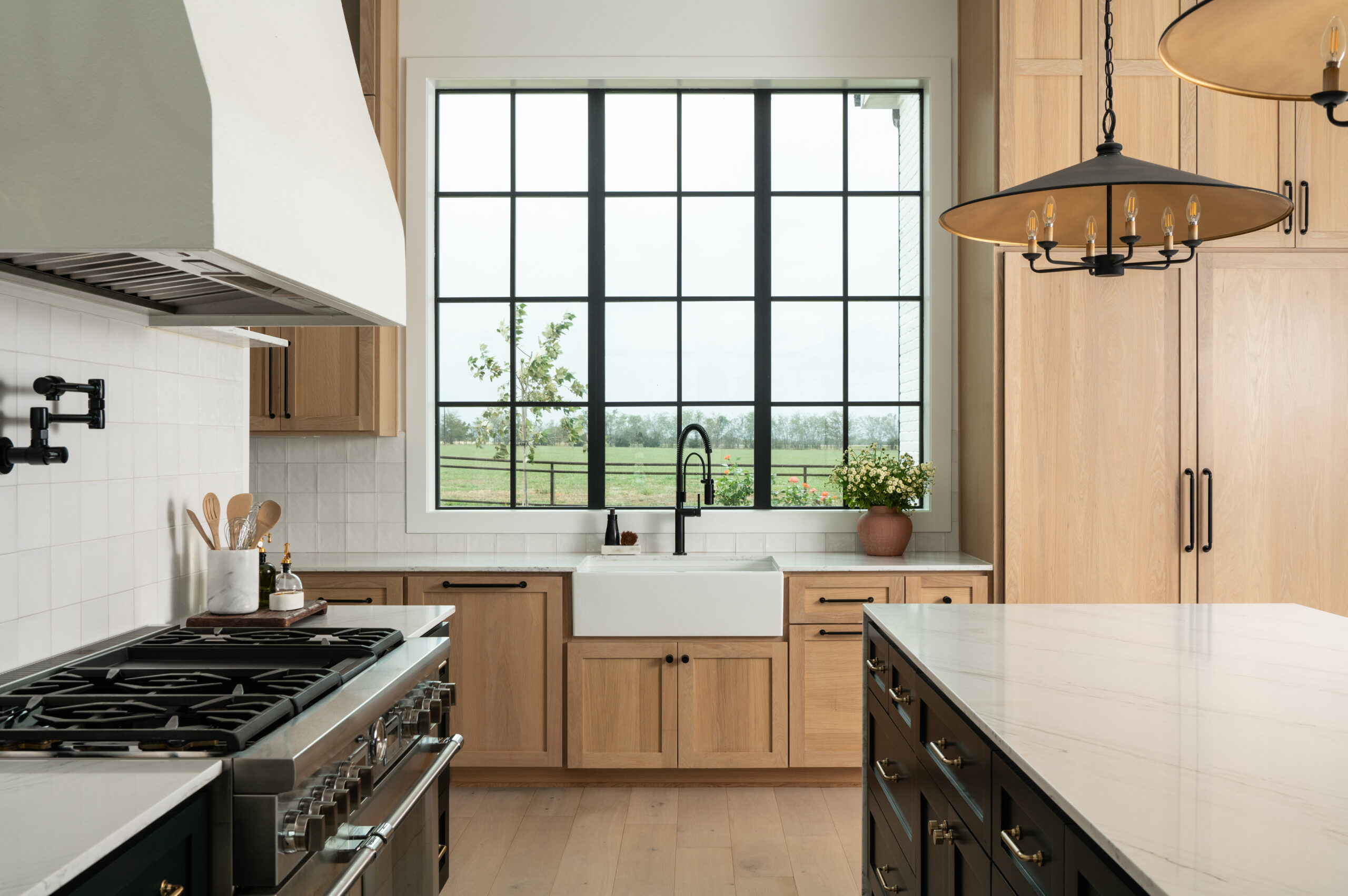 A spacious kitchen with a view of the lush outdoor scenery through a large window above a white farmhouse sink, exemplifying modern home interior design.